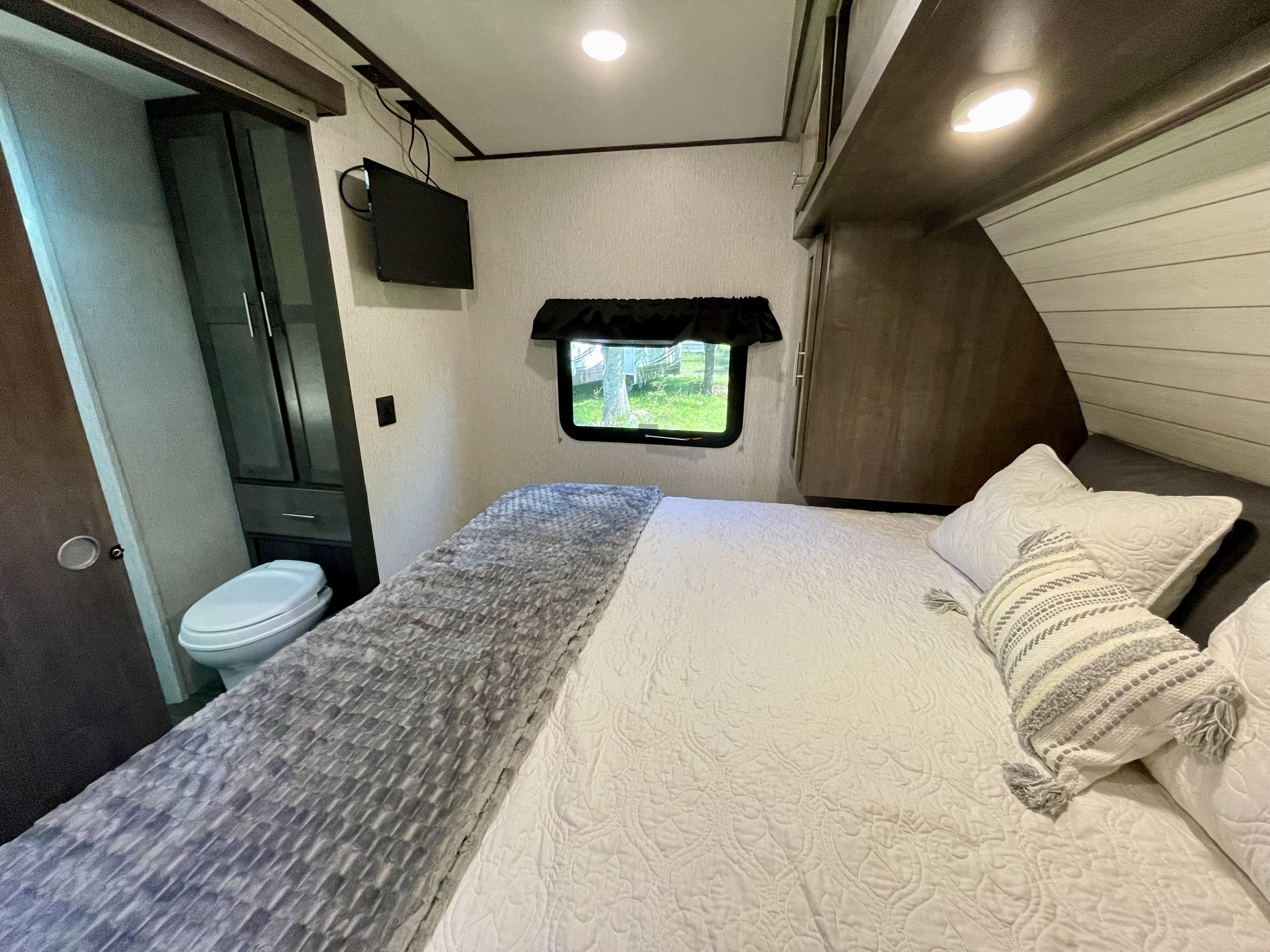 Cozy RV rental bedroom featuring a bed with pillows, window view, wall-mounted TV, and a compact toilet.. Crossroads RV Sunset Trail Super Lite 2019