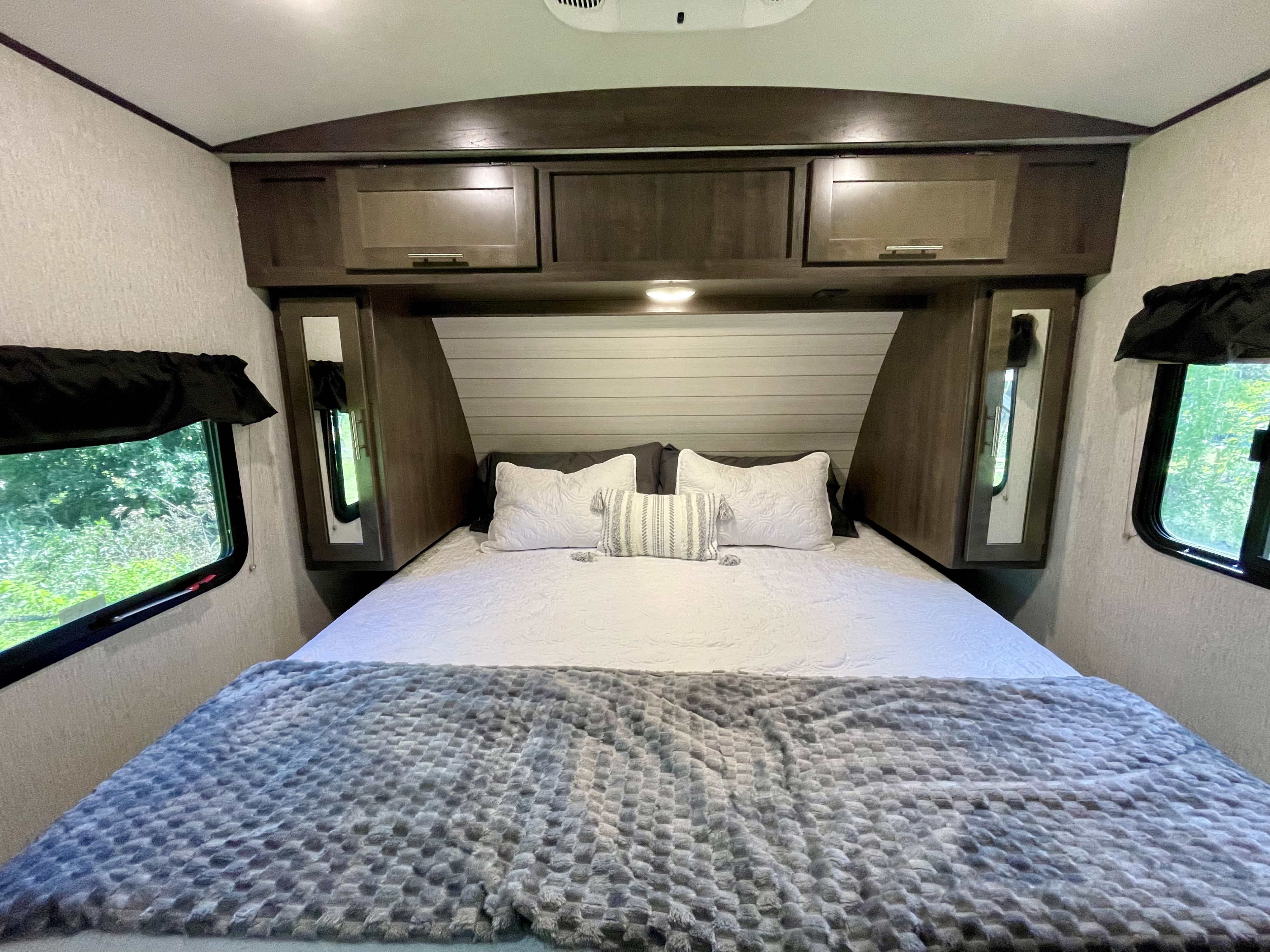 Inviting RV rental bedroom with a neatly made bed, white bedding, cozy gray blanket, overhead cabinets, and side windows.. Crossroads RV Sunset Trail Super Lite 2019