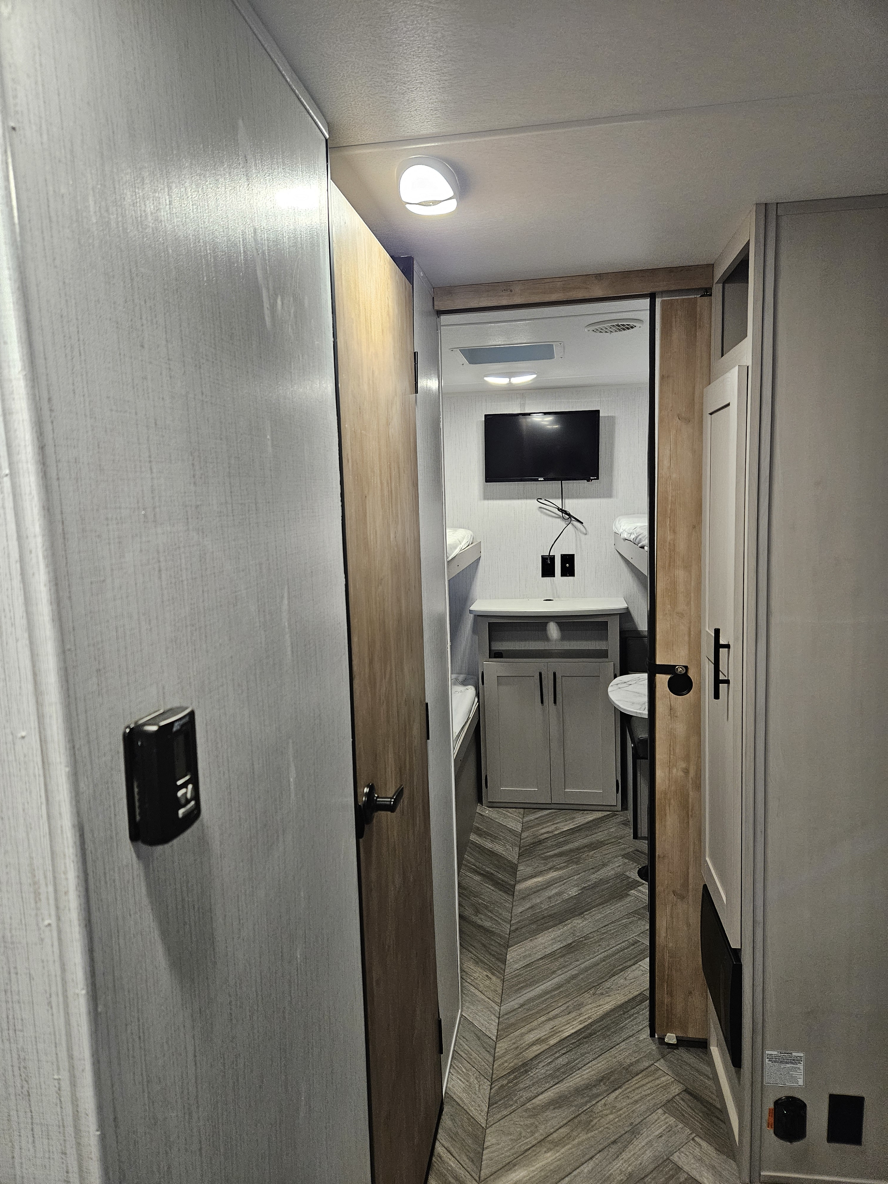 Modern RV rental interior: a hallway leads to bunk beds, a TV, and built-in storage. Chevron floor and ceiling light.. Heartland RVs Prowler 2023