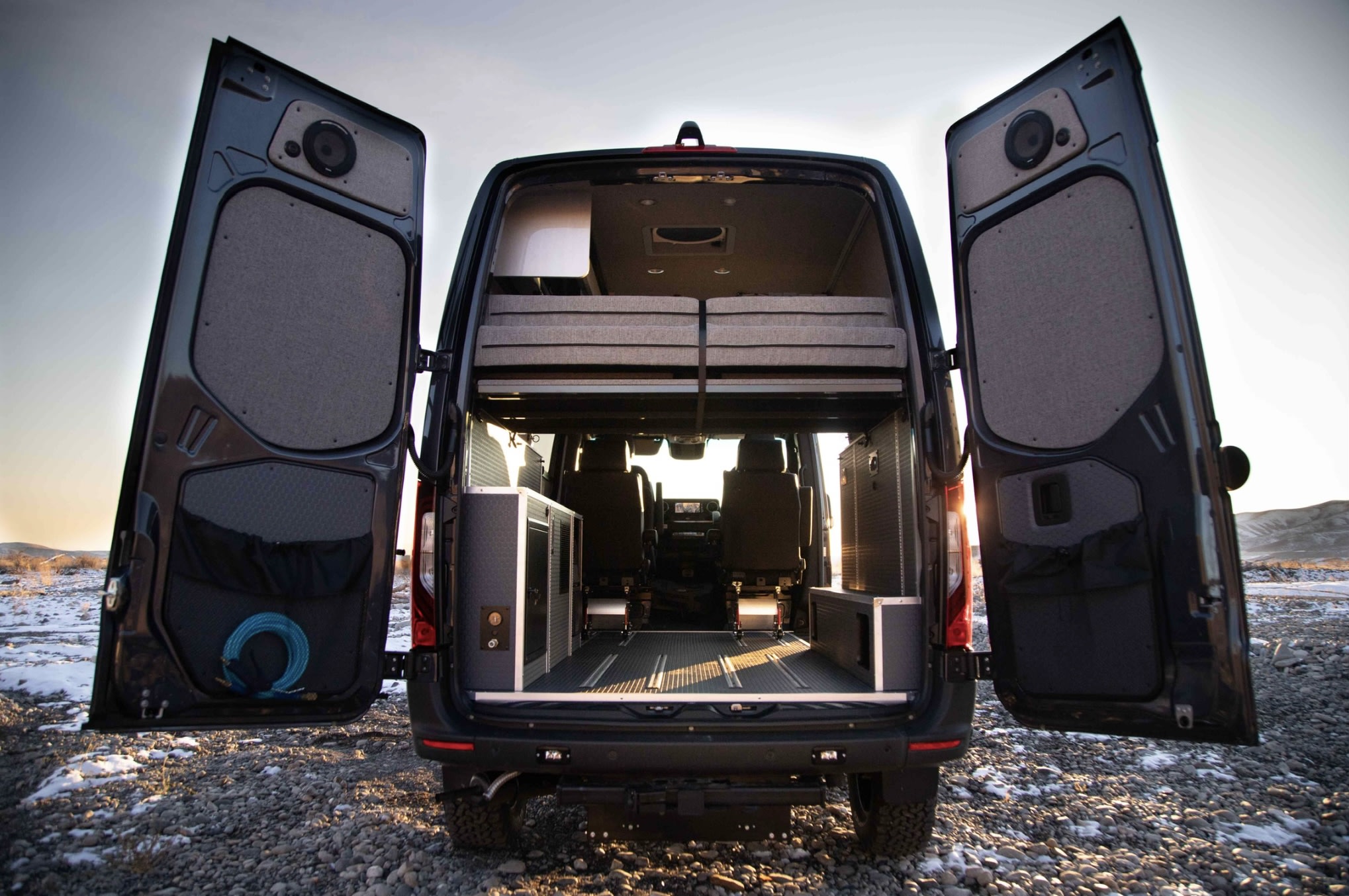 Rear view of a dark camper van with open doors, revealing a bed platform, modular storage, and a durable floor. Ready for RV rental adventures.. Mercedes Sprinter 2020