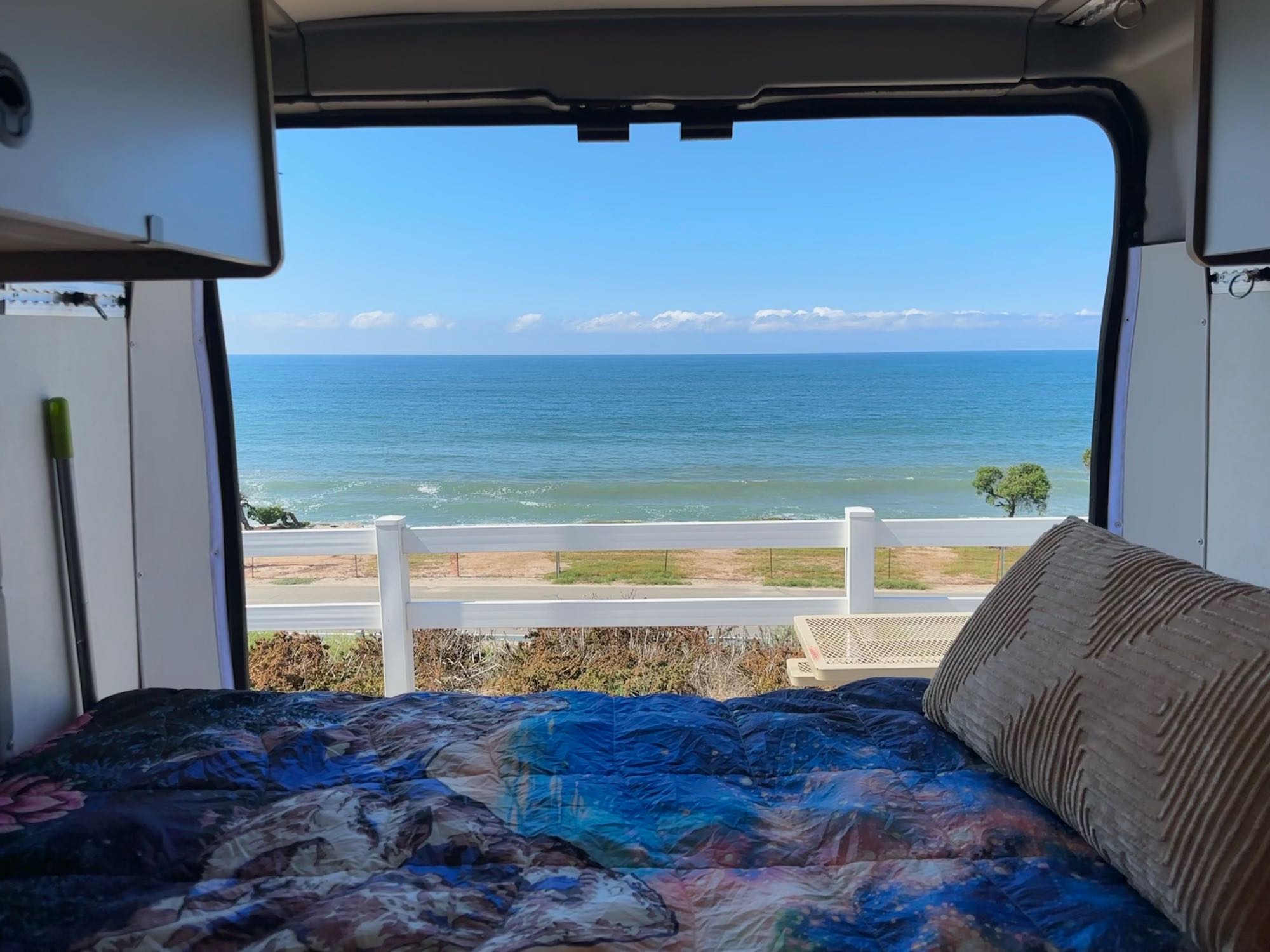 Serene ocean view from an RV rental. Cozy bed inside facing the tranquil sea, clear blue sky, and a white fence below.. Winnebago Solis 2023