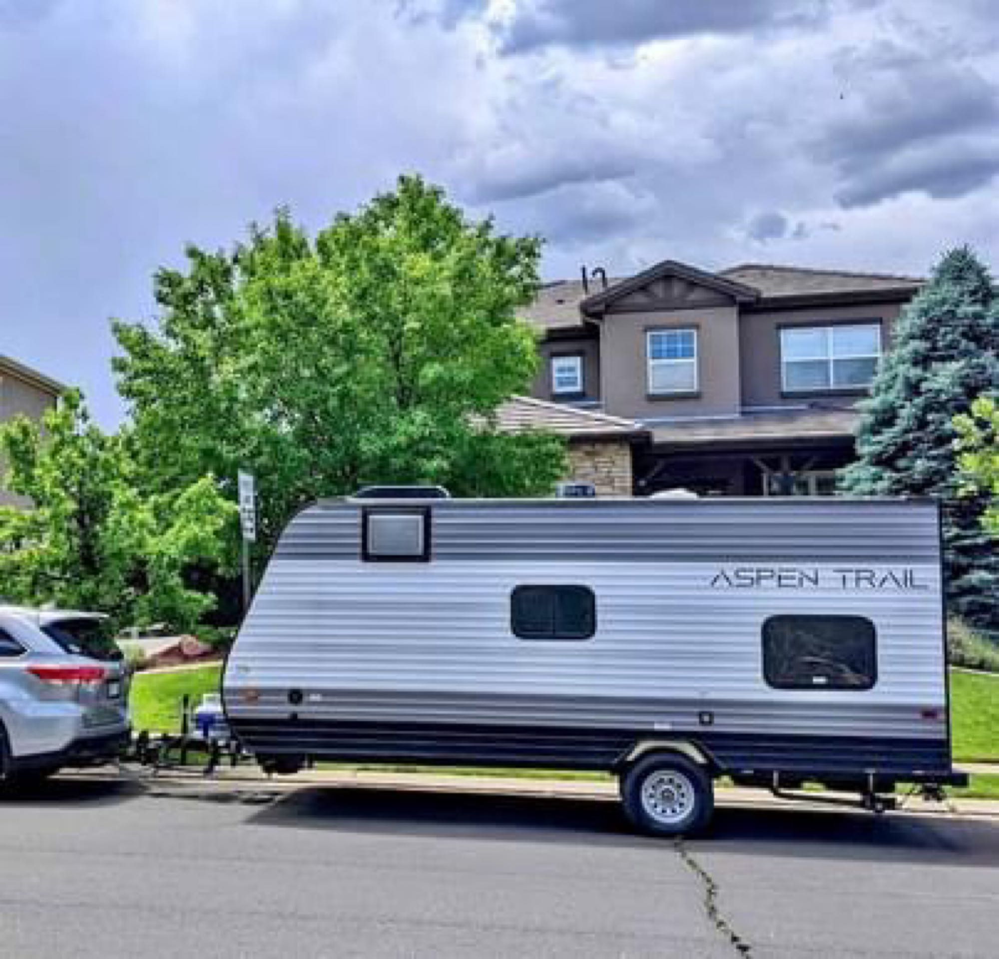 Silver Aspen Trail travel trailer, possibly an rv rental, hitched to an SUV and parked on a street in front of a house.. Aspen Trail LE 2022