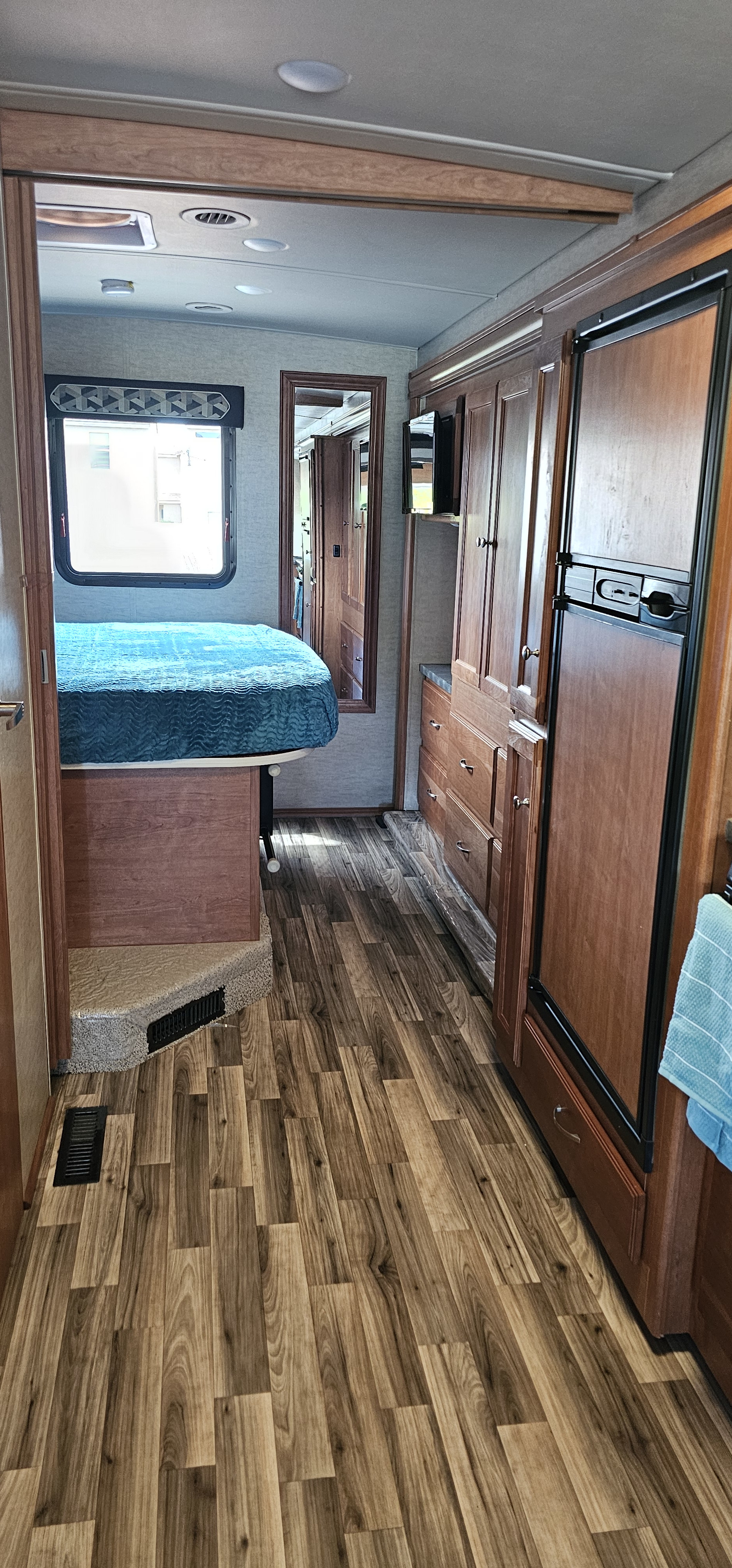 RV rental interior: bedroom with teal bedding, wood-look flooring, and dark wood cabinetry with a fridge and mirror.. Winnebago Vista 2017