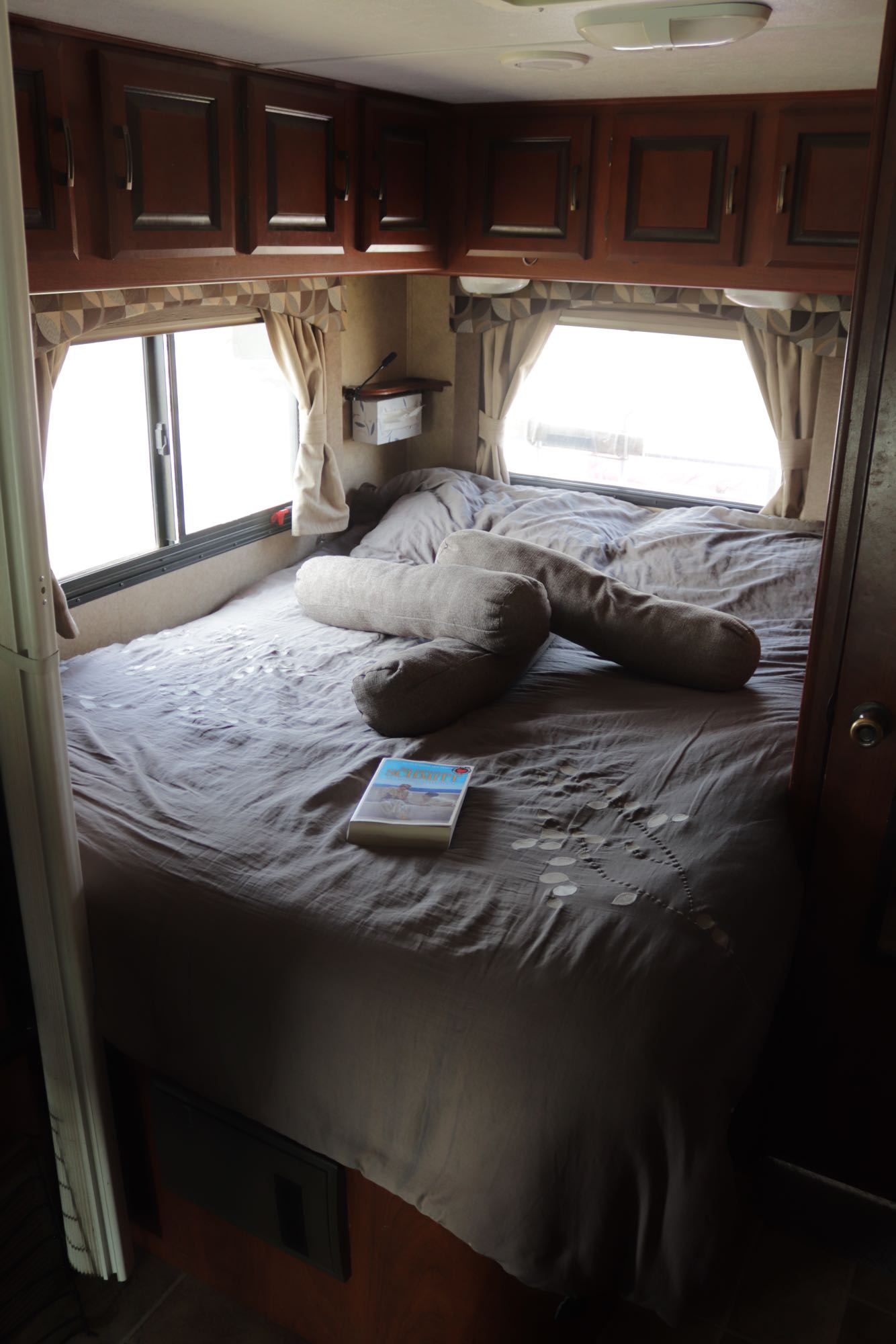 Comfortable RV rental bedroom featuring a grey bed with bolster pillows, windows, and overhead cabinets.. Forest River Sunseeker 2012
