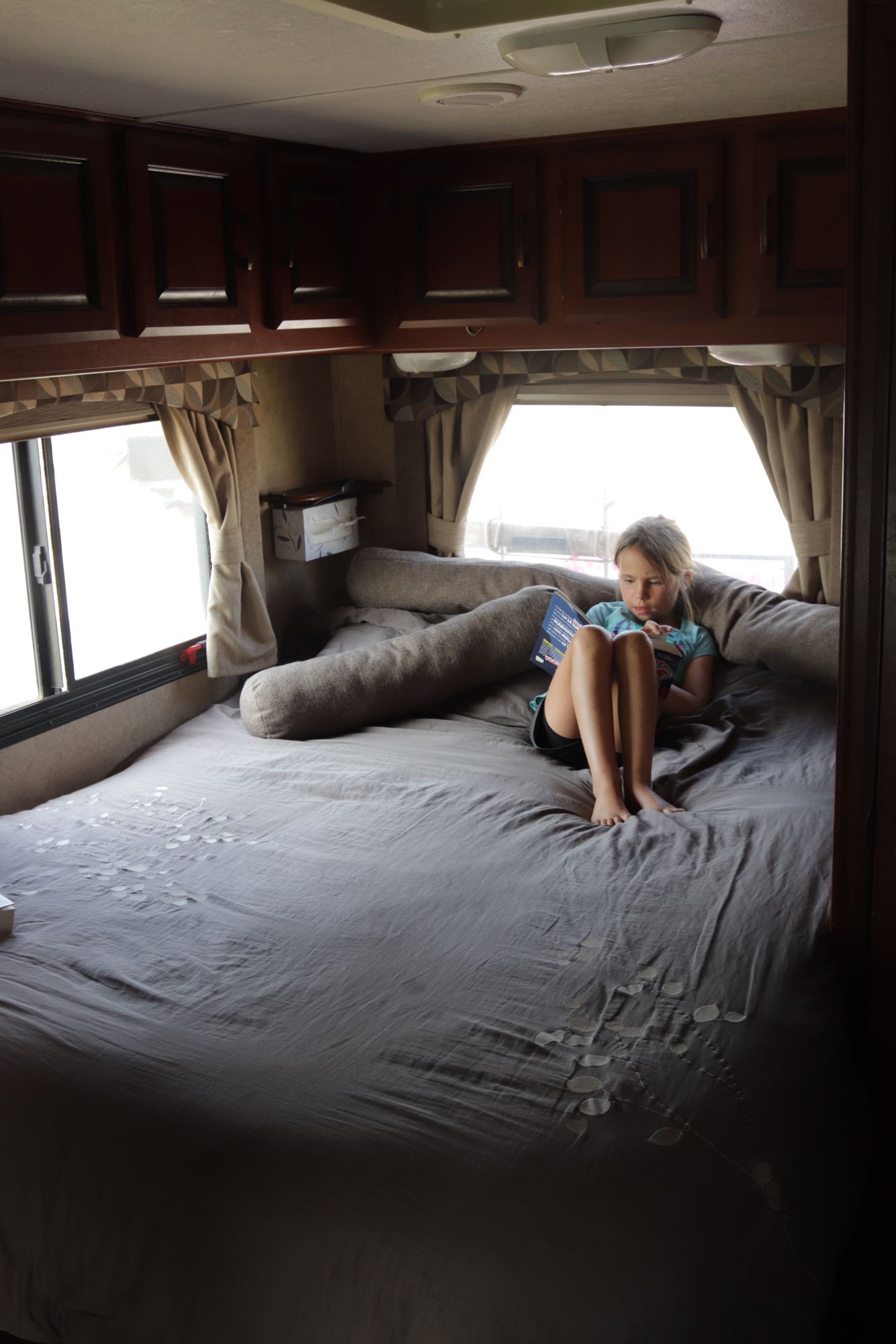 An RV rental provides cozy comfort. A young girl reads a book on a large bed, with windows and overhead storage visible.. Forest River Sunseeker 2012