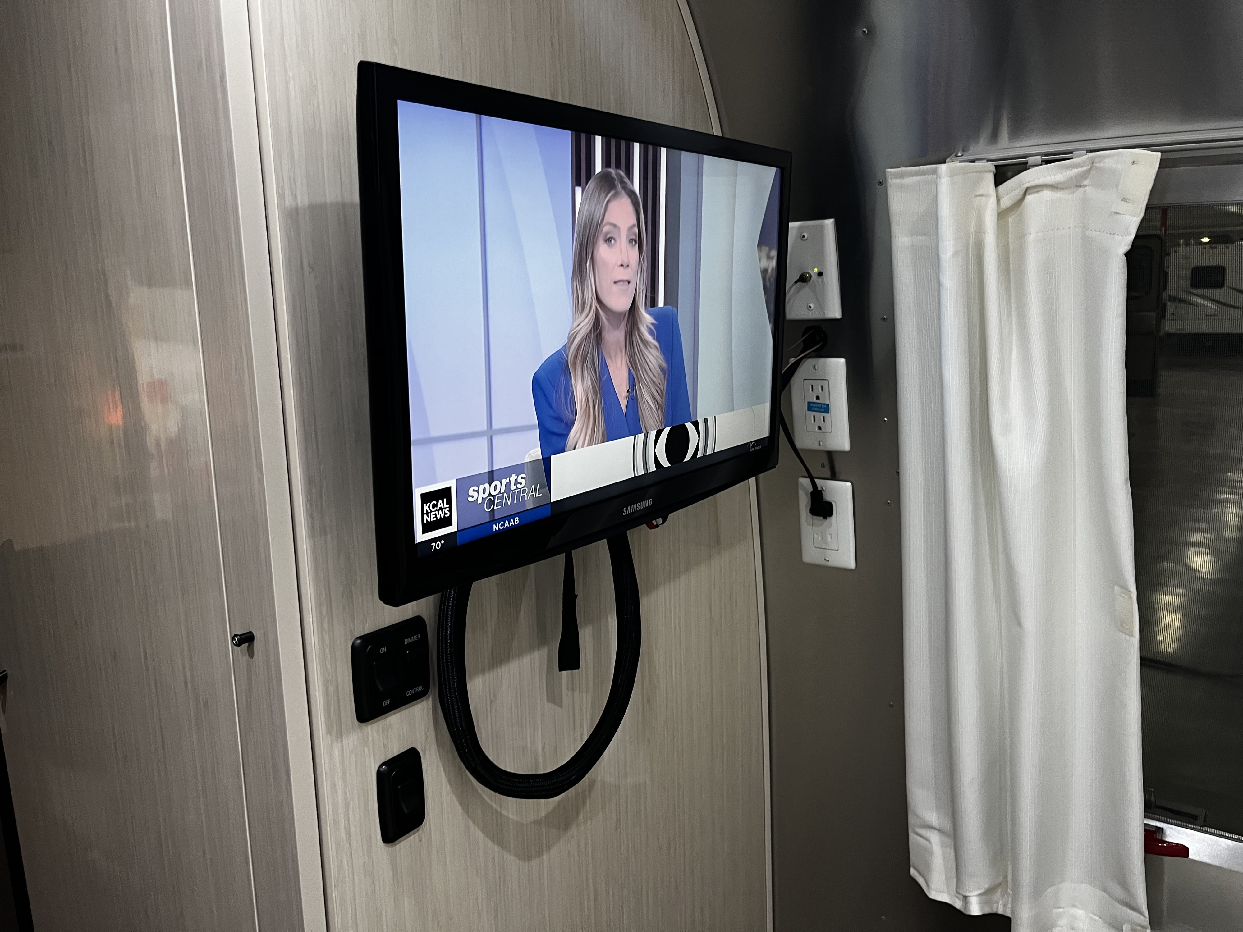View inside a sleek rv rental, featuring a wall-mounted TV playing news, with power outlets and a privacy curtain.. Airstream International 2017