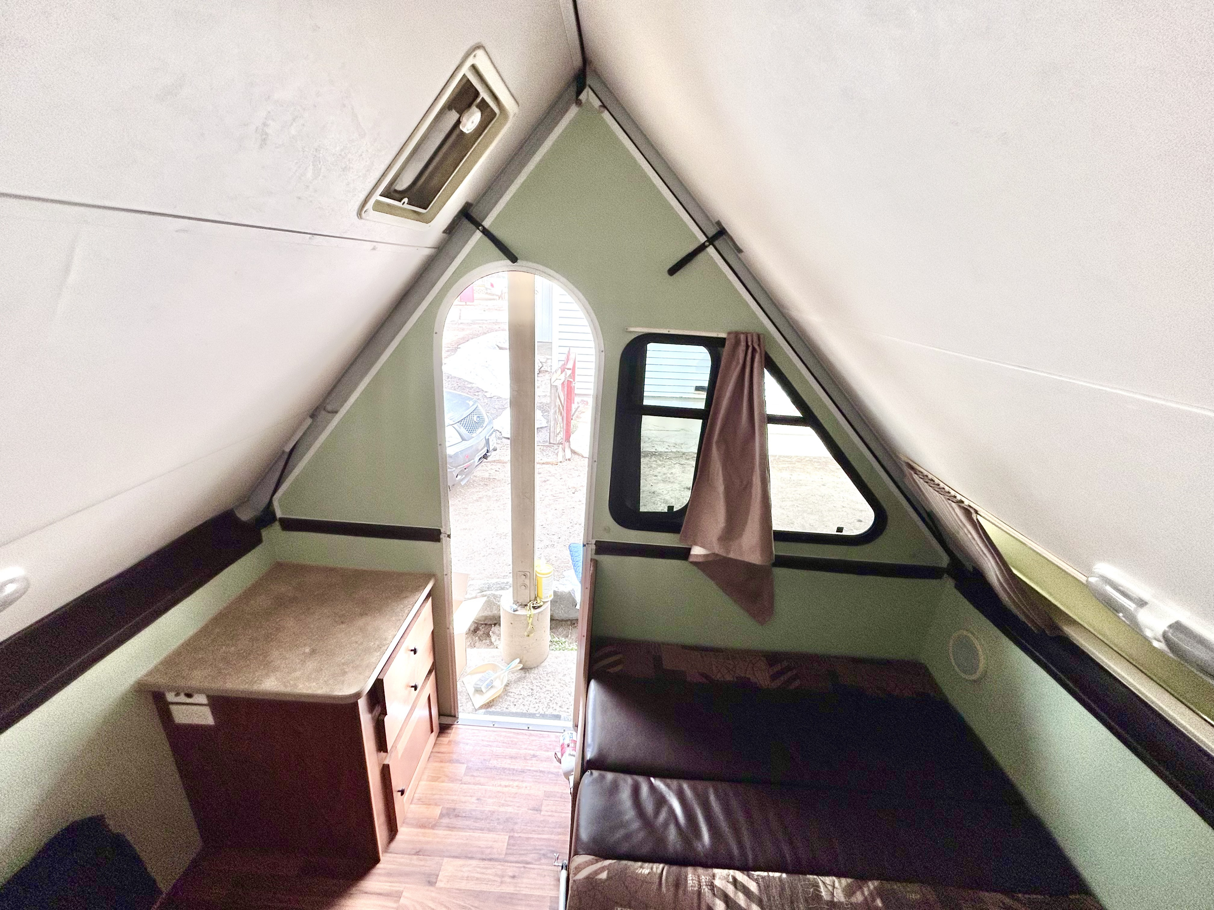 Inside a compact A-frame camper, showing the bed, counter, and open doorway to the outside. A cozy RV rental option.. Chalet Chalet Popup Trailer 2013