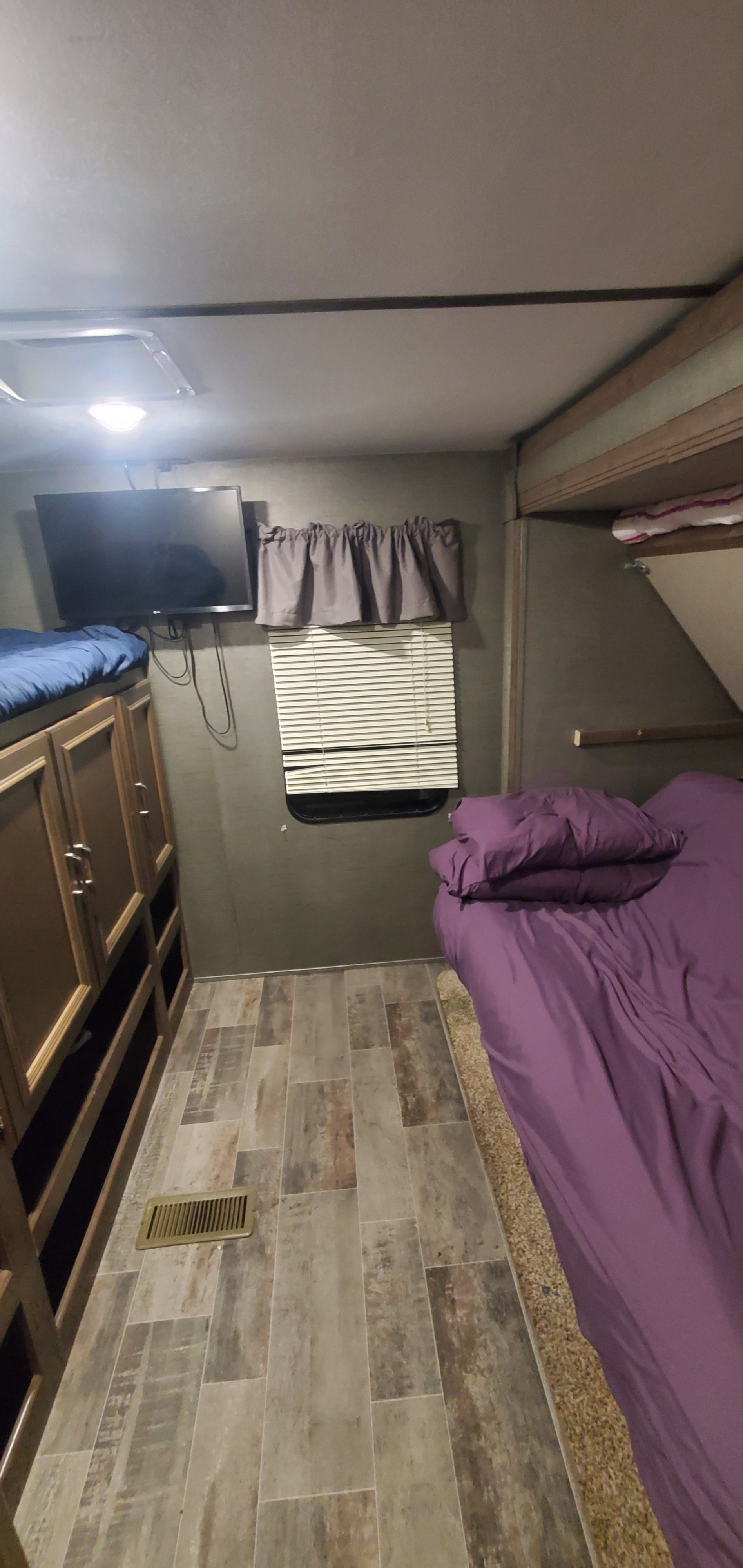 Cozy RV rental bedroom featuring bunk beds, a TV, and a main bed with purple bedding. Includes storage cabinets and a window.. Keystone RV Passport Grand Touring 2019