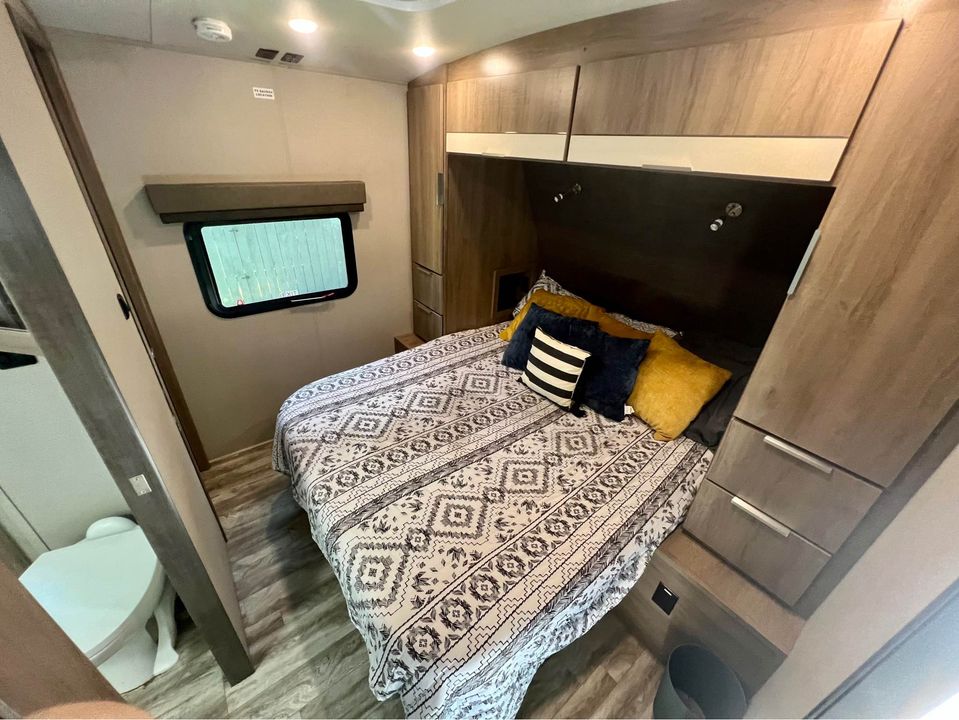 Inside an RV rental: a compact bedroom featuring a bed, storage cabinets, window, and nearby toilet. Ready for your road trip.. Grand Design Imagine 2020