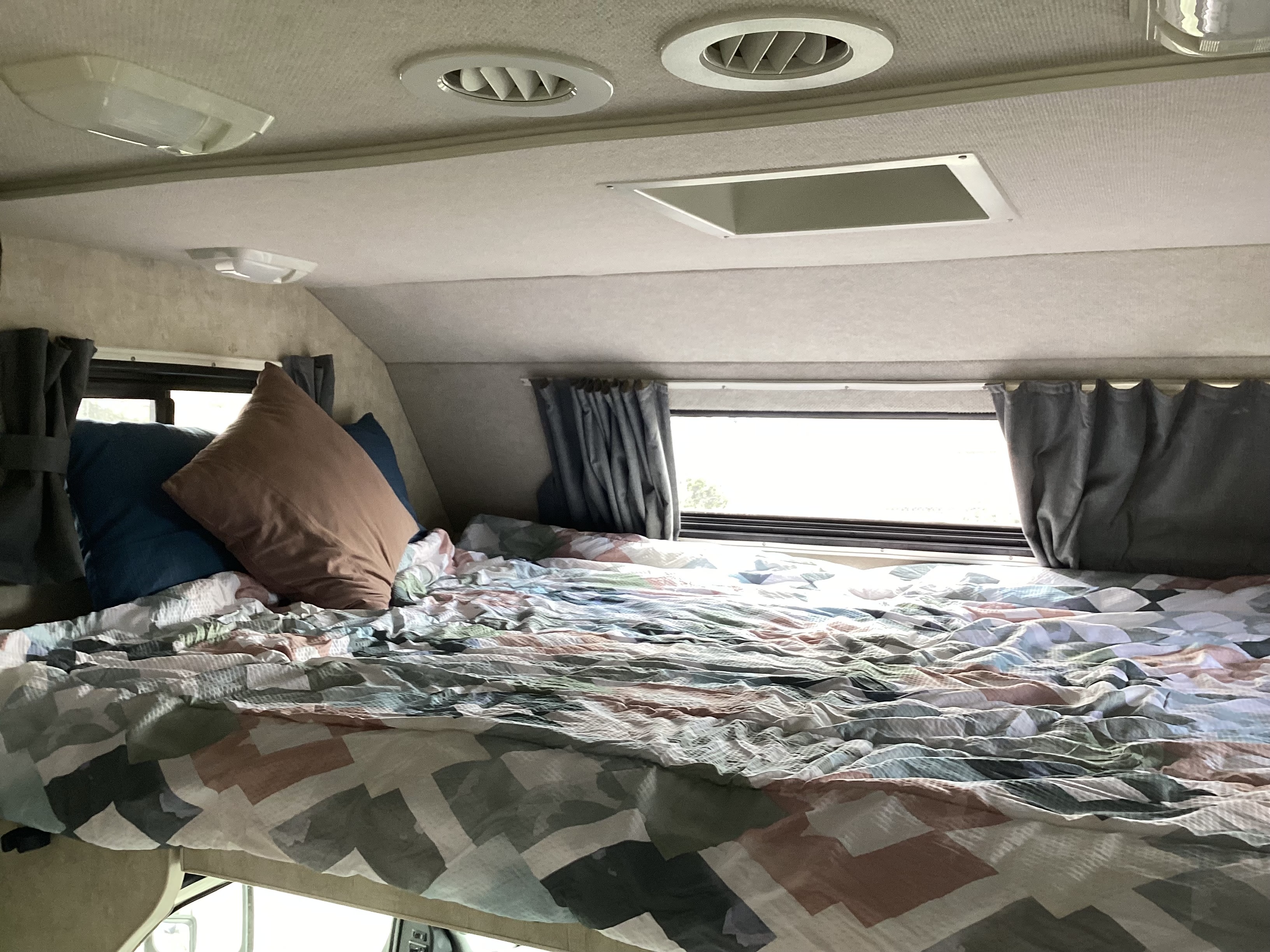View of a cozy RV rental sleeping nook featuring a patterned bed, pillows, windows with curtains, and ceiling fixtures.. Jamboree Jamboree Motorhome 2005