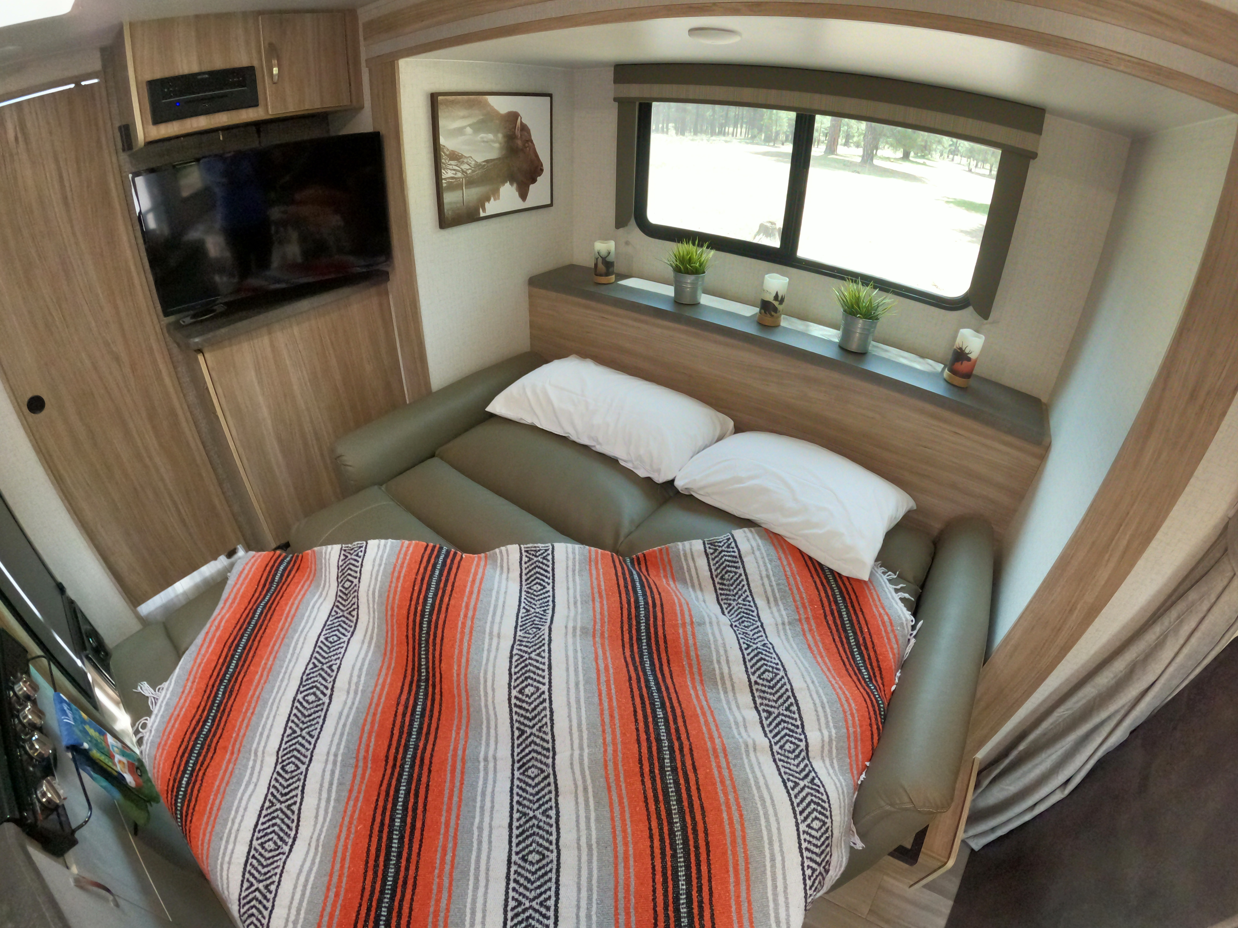 Comfortable rv rental bedroom with a striped blanket, wall-mounted TV, and window showcasing a peaceful outdoor forest.. Winnebago Micro Minnie 2022