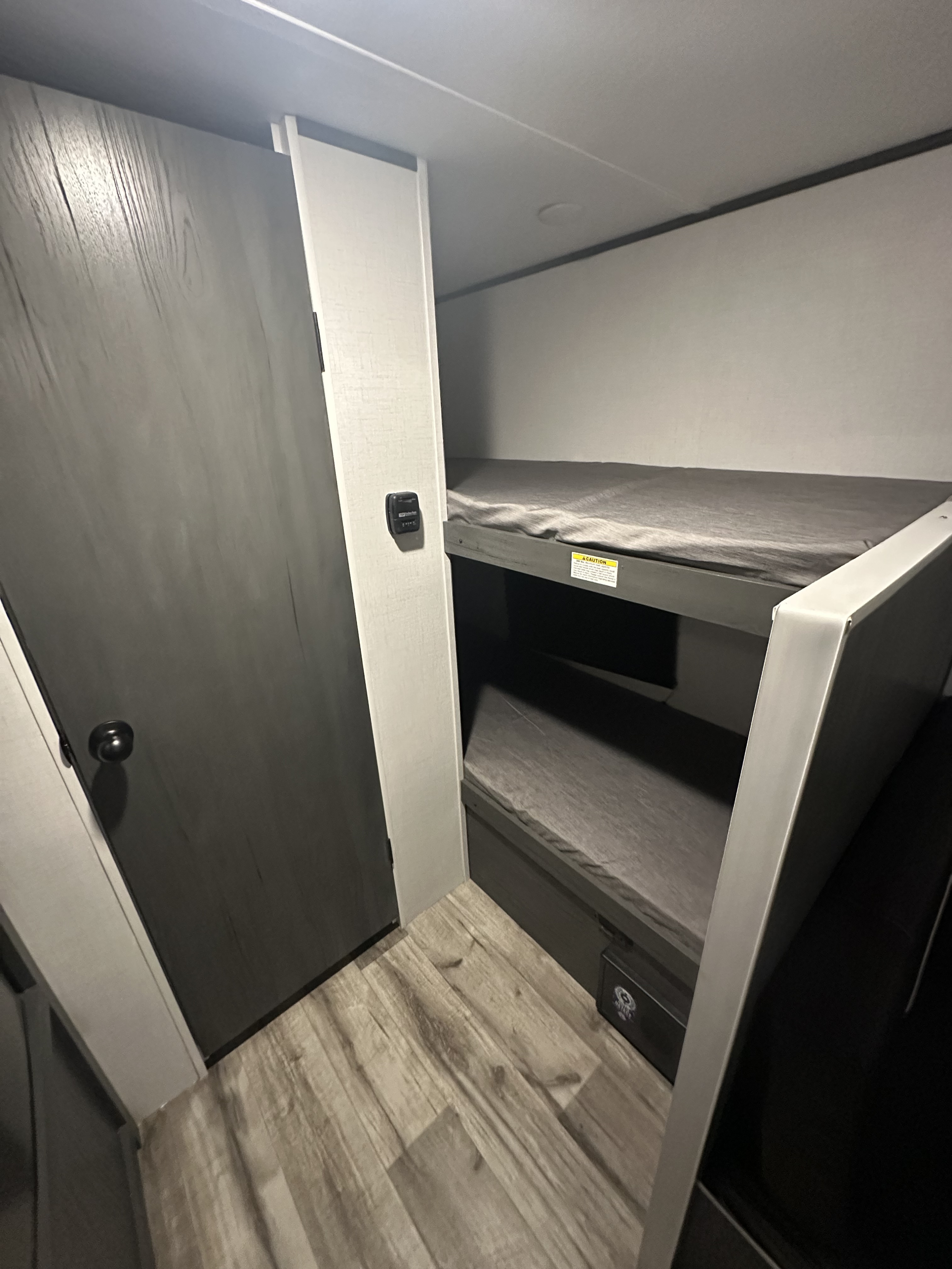 Explore this RV rental's cozy bunk area: two stacked beds with dark grey linens, a textured grey door, and light wood flooring.. Dutchmen Colorado 2024