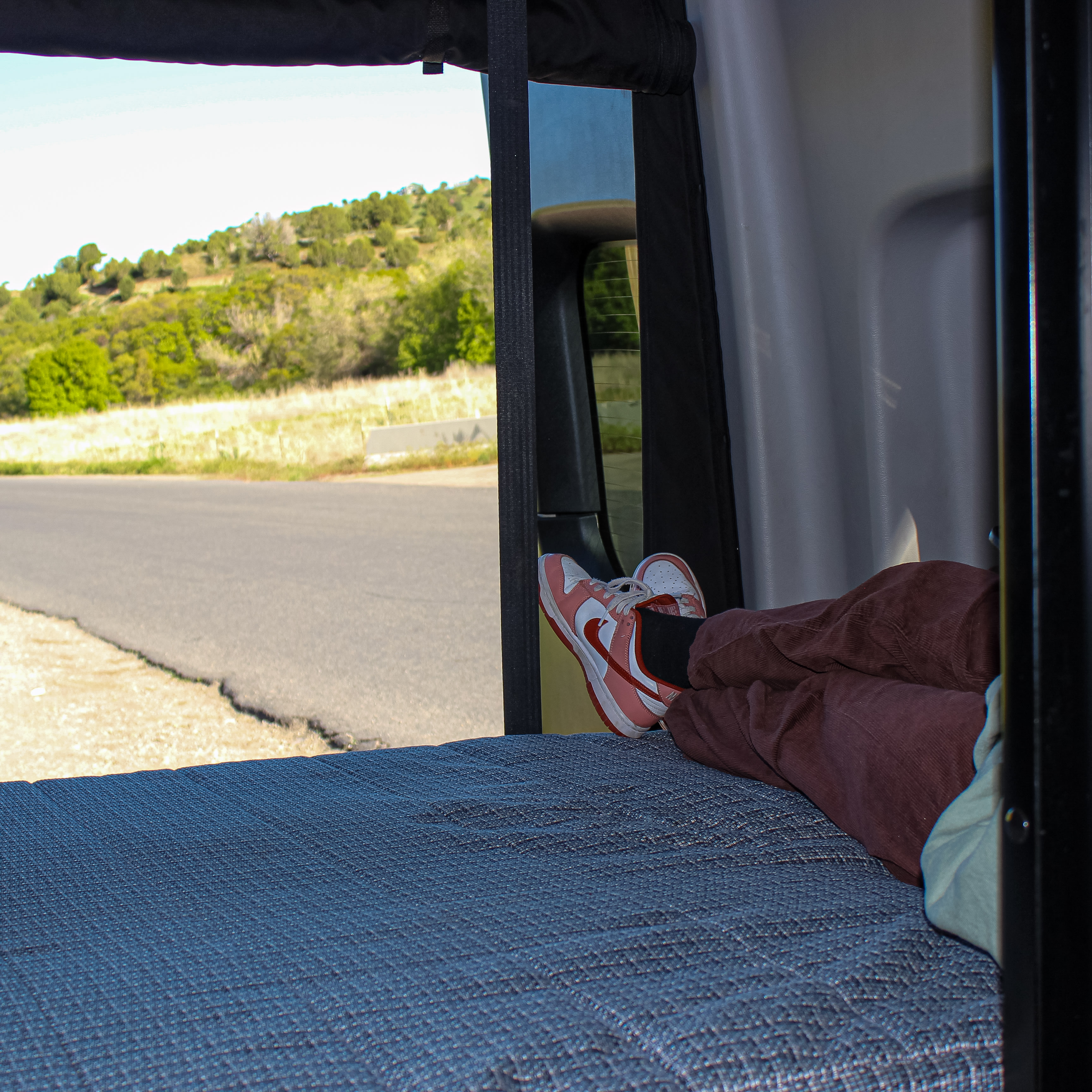 Cozy RV rental comfort: feet up on the bed, enjoying the scenic road trip view outside.. Mercedes-Benz Sprinter 2021