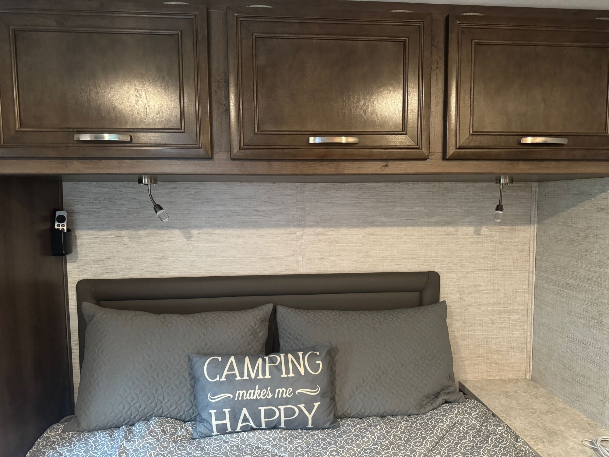 An RV rental bedroom featuring overhead cabinets, reading lights, a comfy bed, and a "Camping makes me HAPPY" pillow.. Jayco Redhawk 2021