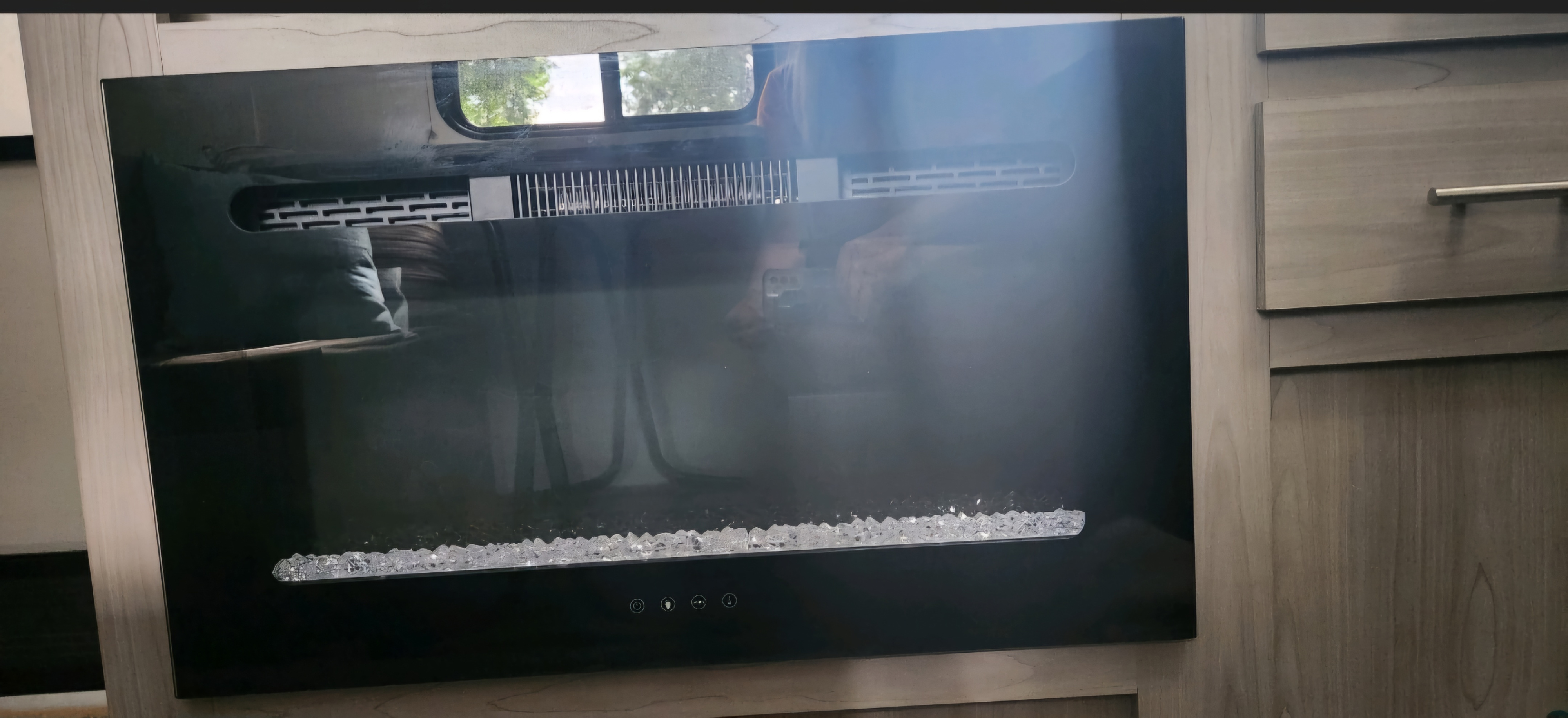 An electric fireplace unit with crystal features and control buttons, built into the wood cabinetry of an RV rental.. Dutchmen Coleman Lantern 2023