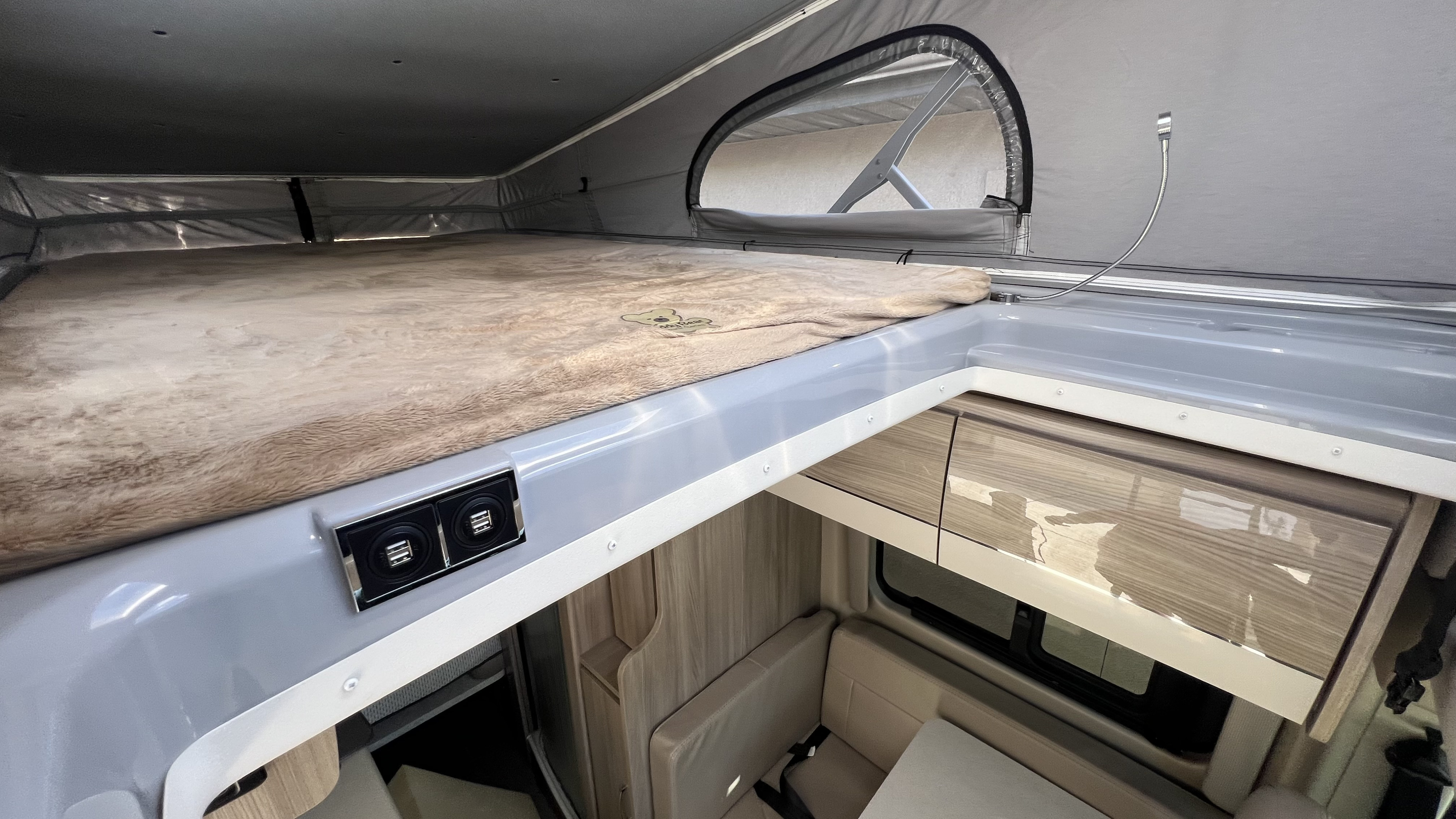 Inside an RV rental, showing the comfy pop-top bed with USB ports above the lower living area and cabinets.. Thor Sequnce 2023