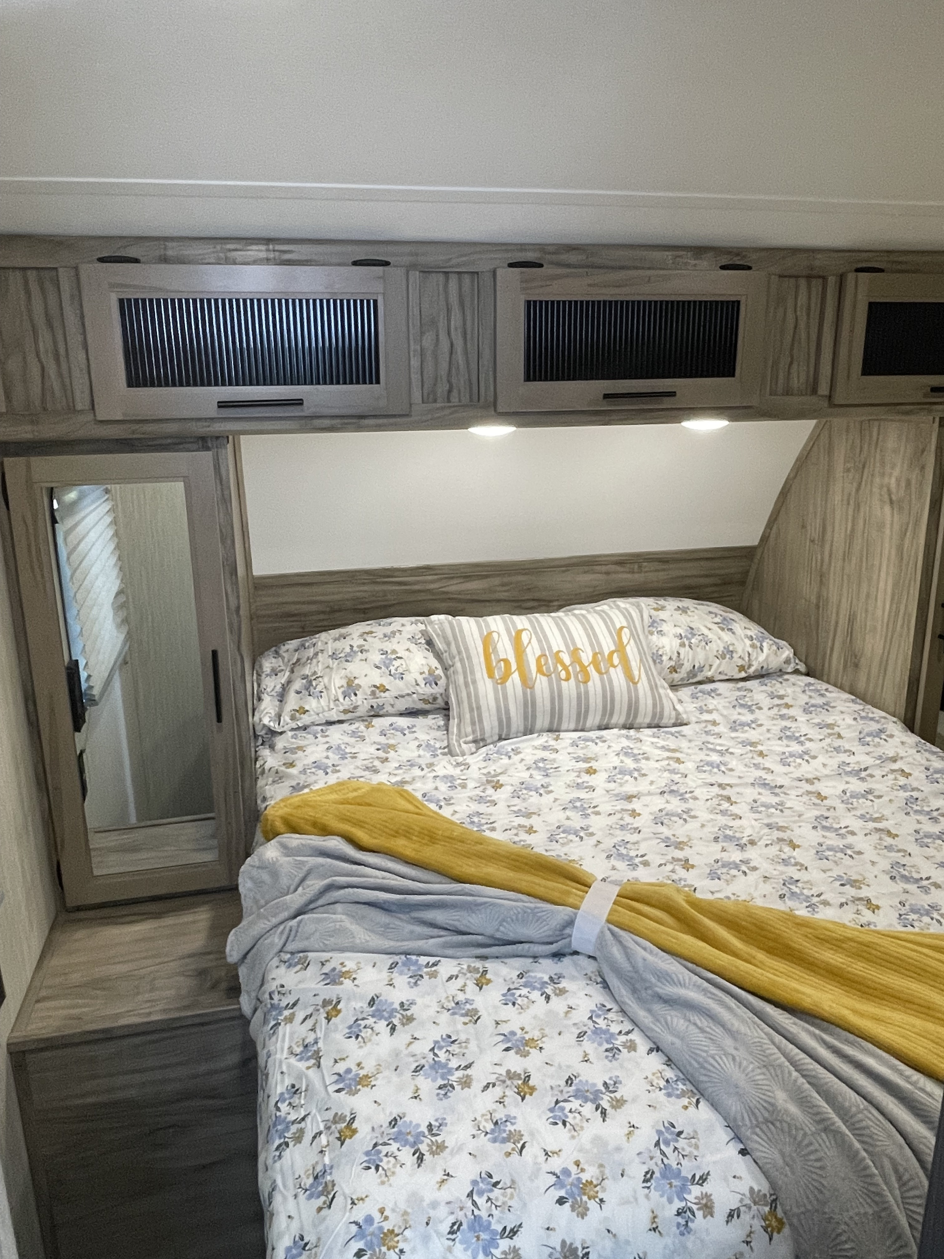 Cozy RV rental bedroom features floral bedding, a 'blessed' pillow, overhead cabinets, and a full-length mirror, perfect for travelers.. Forest River Cherokee Alpha Wolf 2022