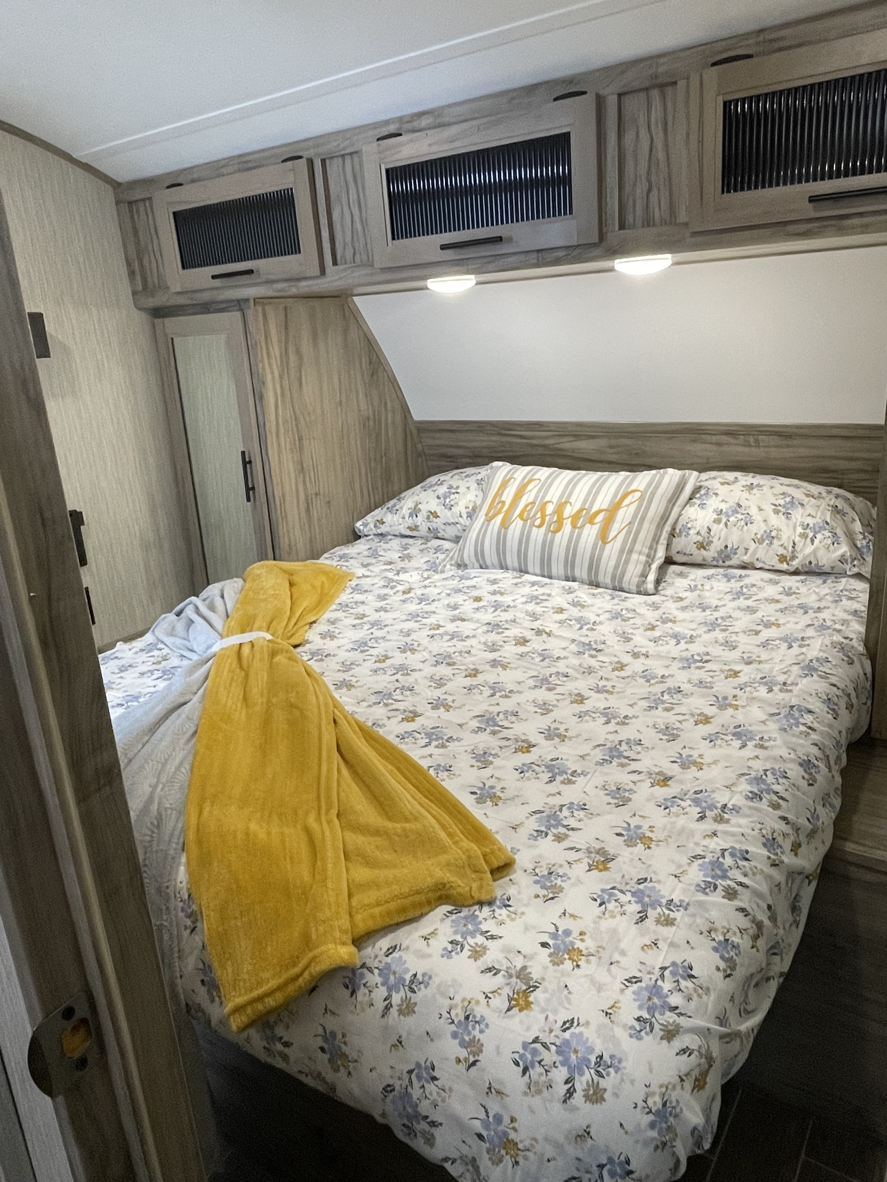 Cozy RV rental bedroom with floral bedding, a yellow blanket, and 'Blessed' pillow, featuring overhead storage cabinets.. Forest River Cherokee Alpha Wolf 2022