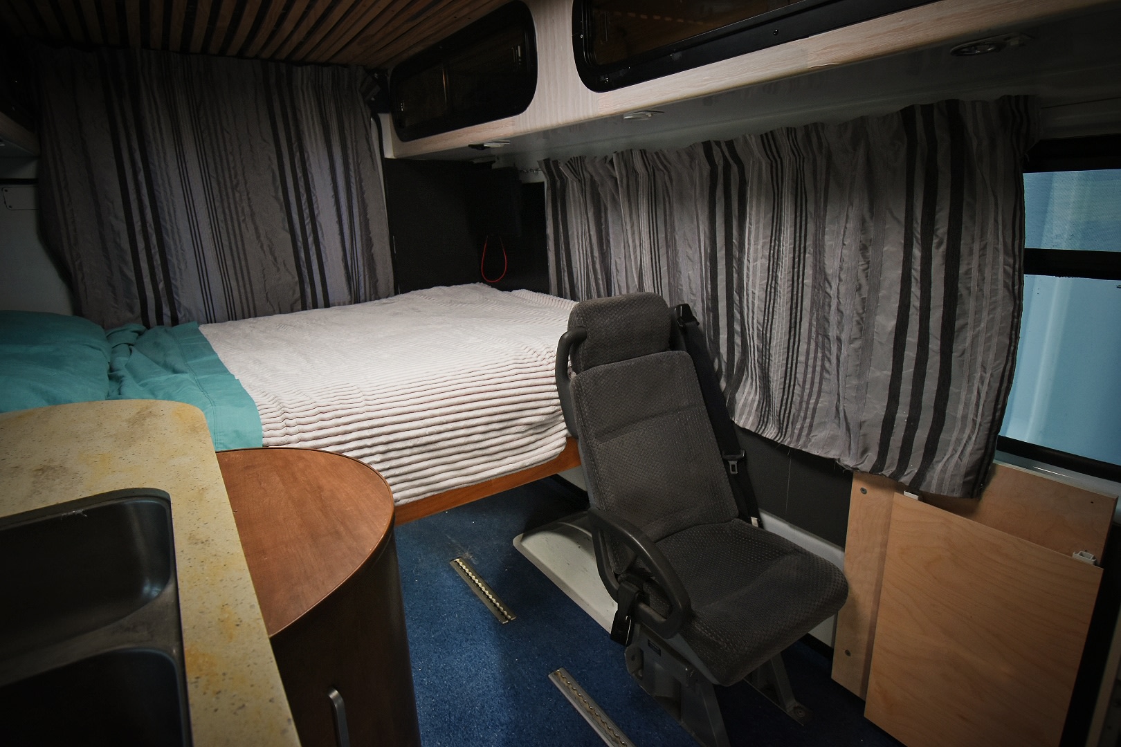 Inside a cozy rv rental, showing a comfortable bed, sink, passenger seat, and striped curtains. Wooden ceiling above blue carpet.. Mercedes-Benz Sprinter 2006