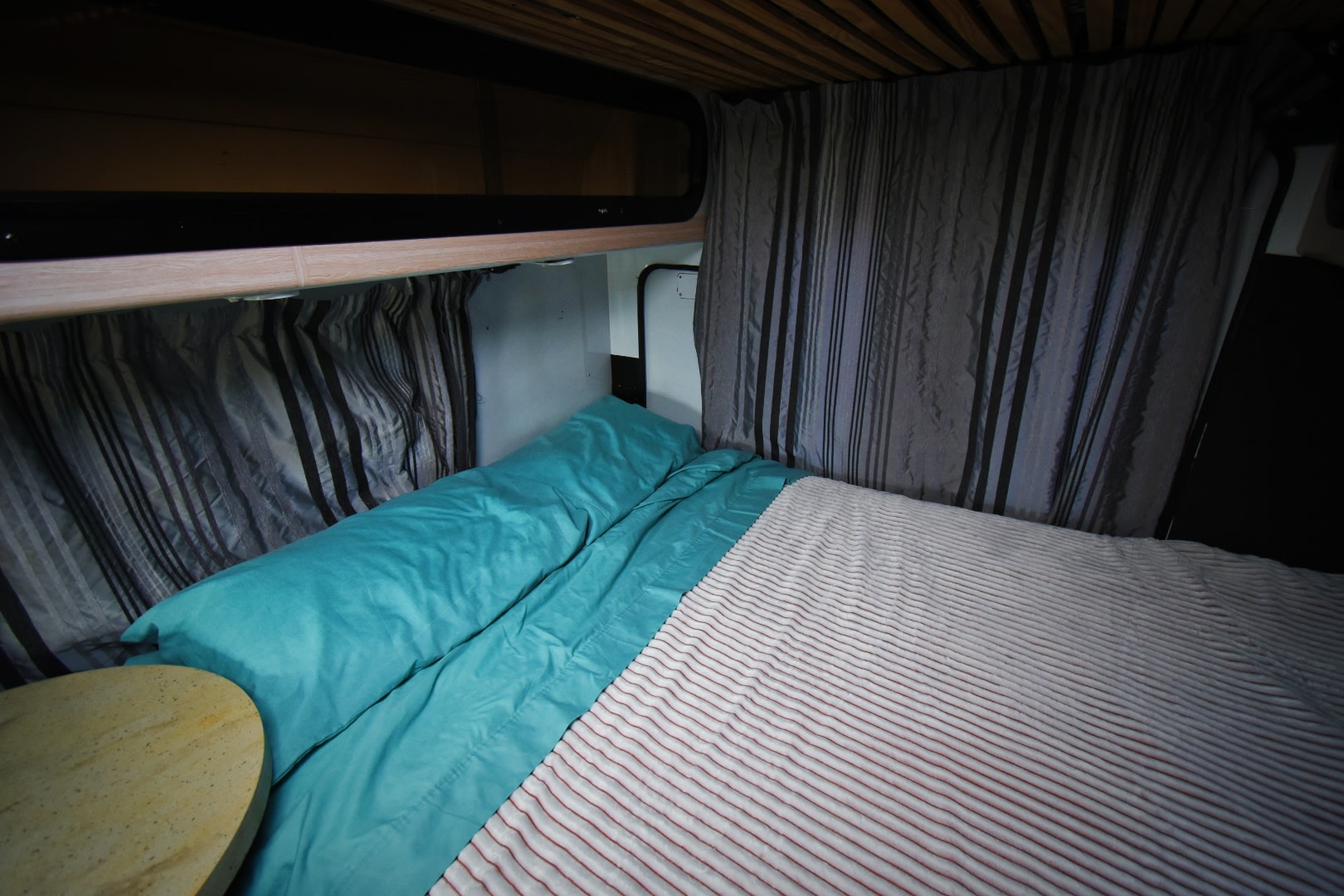 Cozy RV rental bed featuring a teal duvet, striped blanket, and privacy curtains inside a camper.. Mercedes-Benz Sprinter 2006