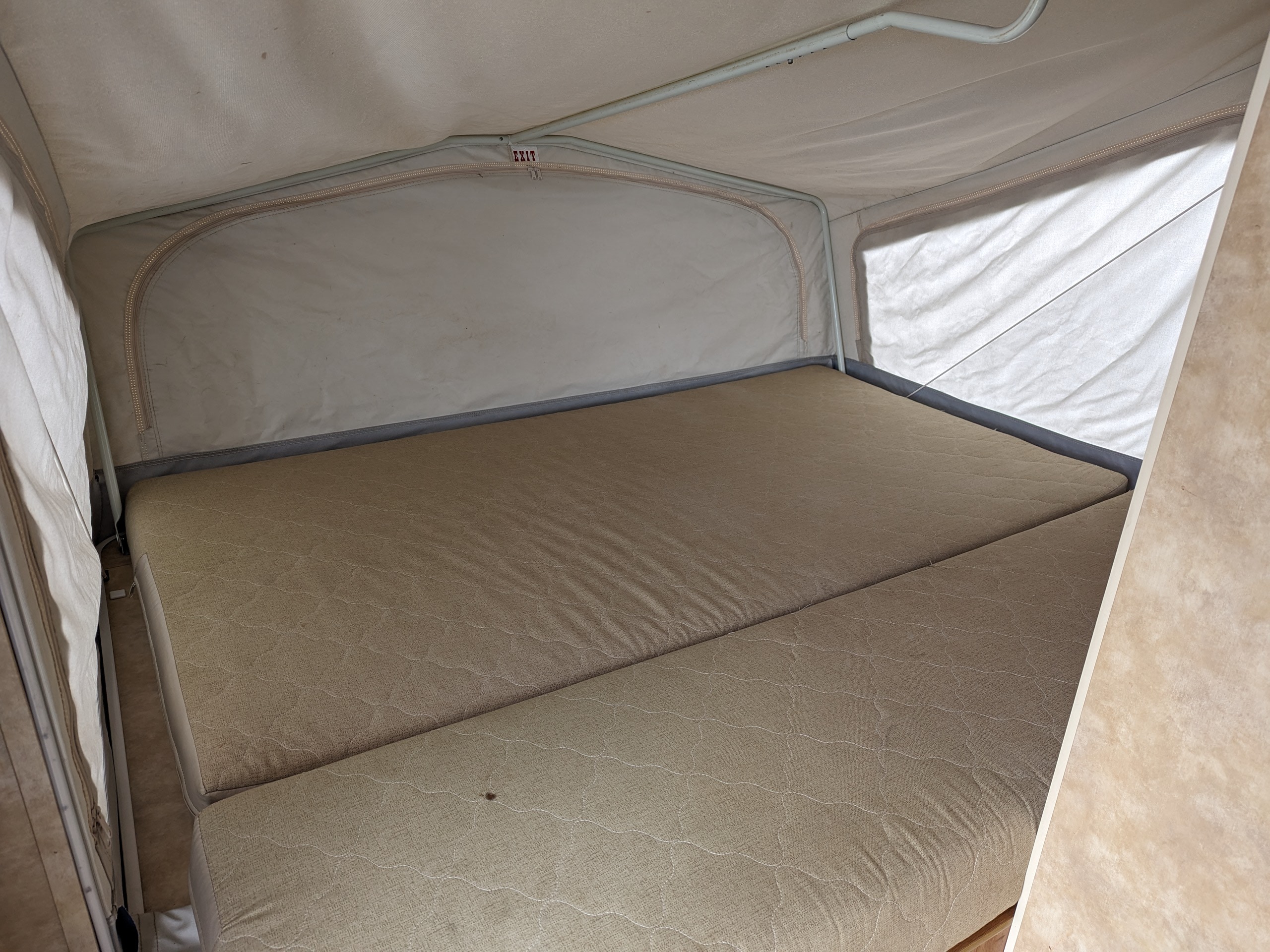 Cozy pop-up bed inside an rv rental, featuring a segmented mattress, light fabric walls, and an 'EXIT' sign overhead.. Jayco Ultralite 2008
