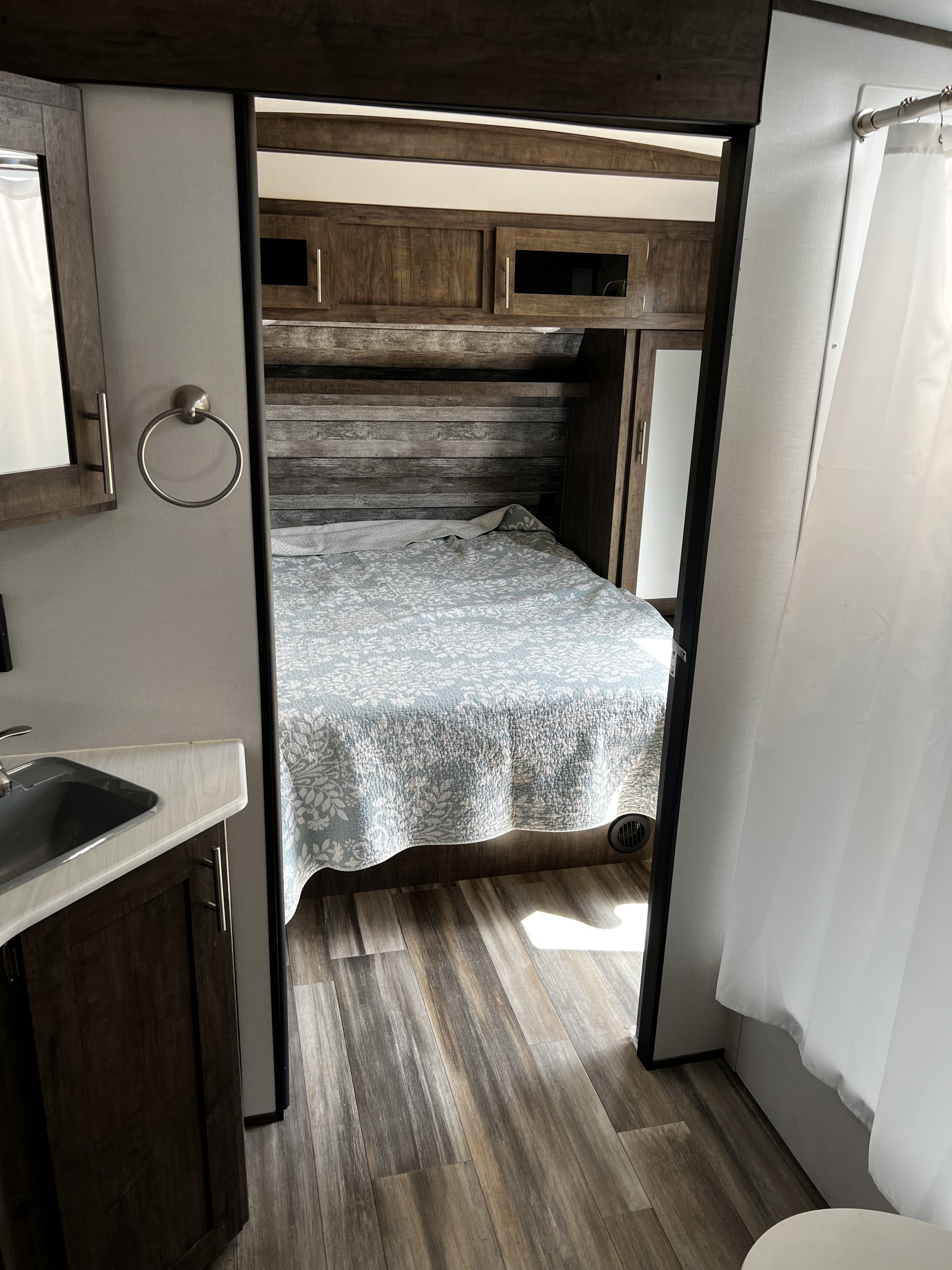 Explore a compact RV rental interior: bedroom with wood headboard, sink vanity, and partial shower view.. Riverside RV Intrepid 2021