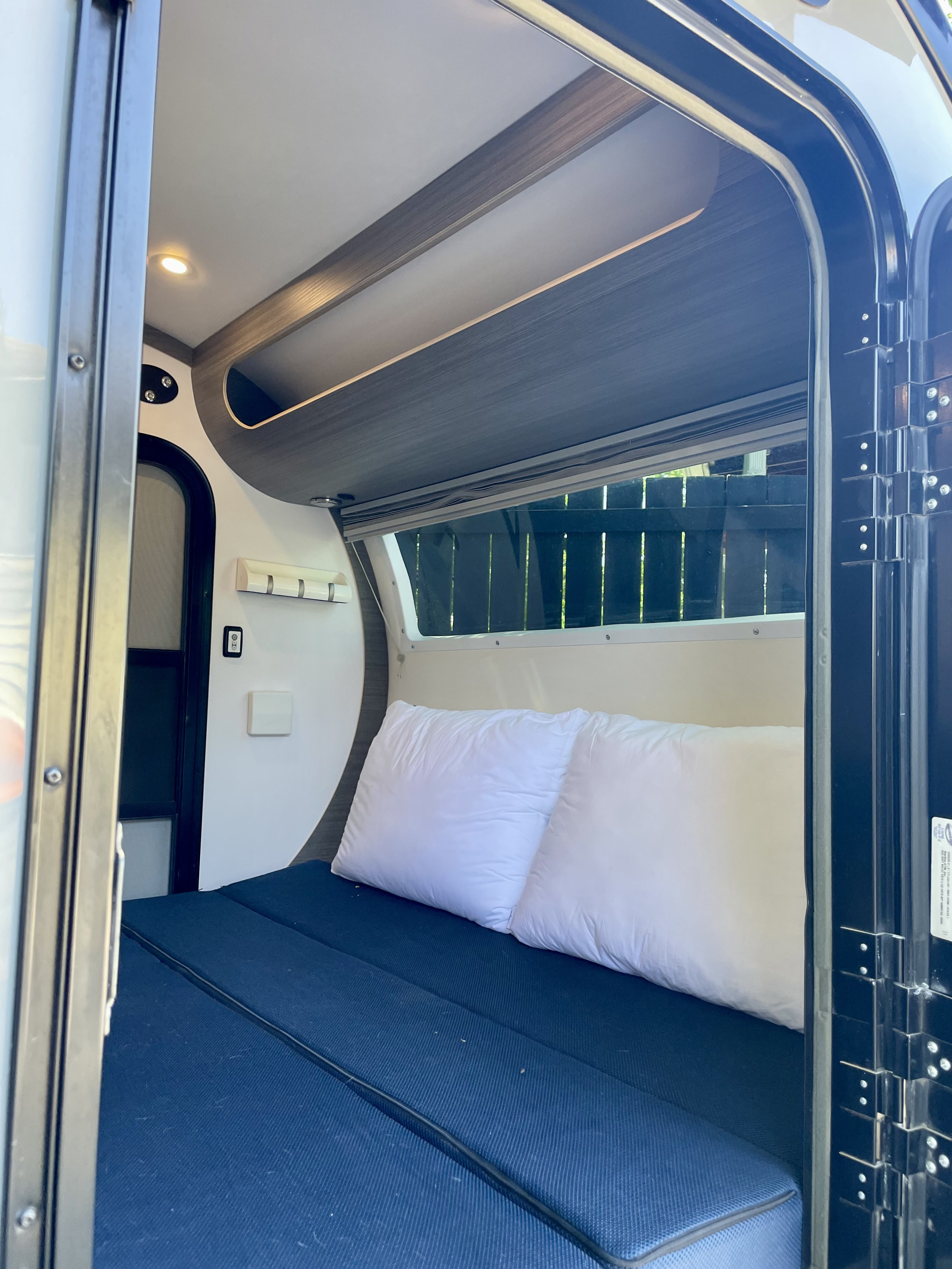 Bright RV rental interior with a blue bed, white pillows, overhead storage, and a window view to a fence.. Bean Trailer Bean Trailer Trailer 2020