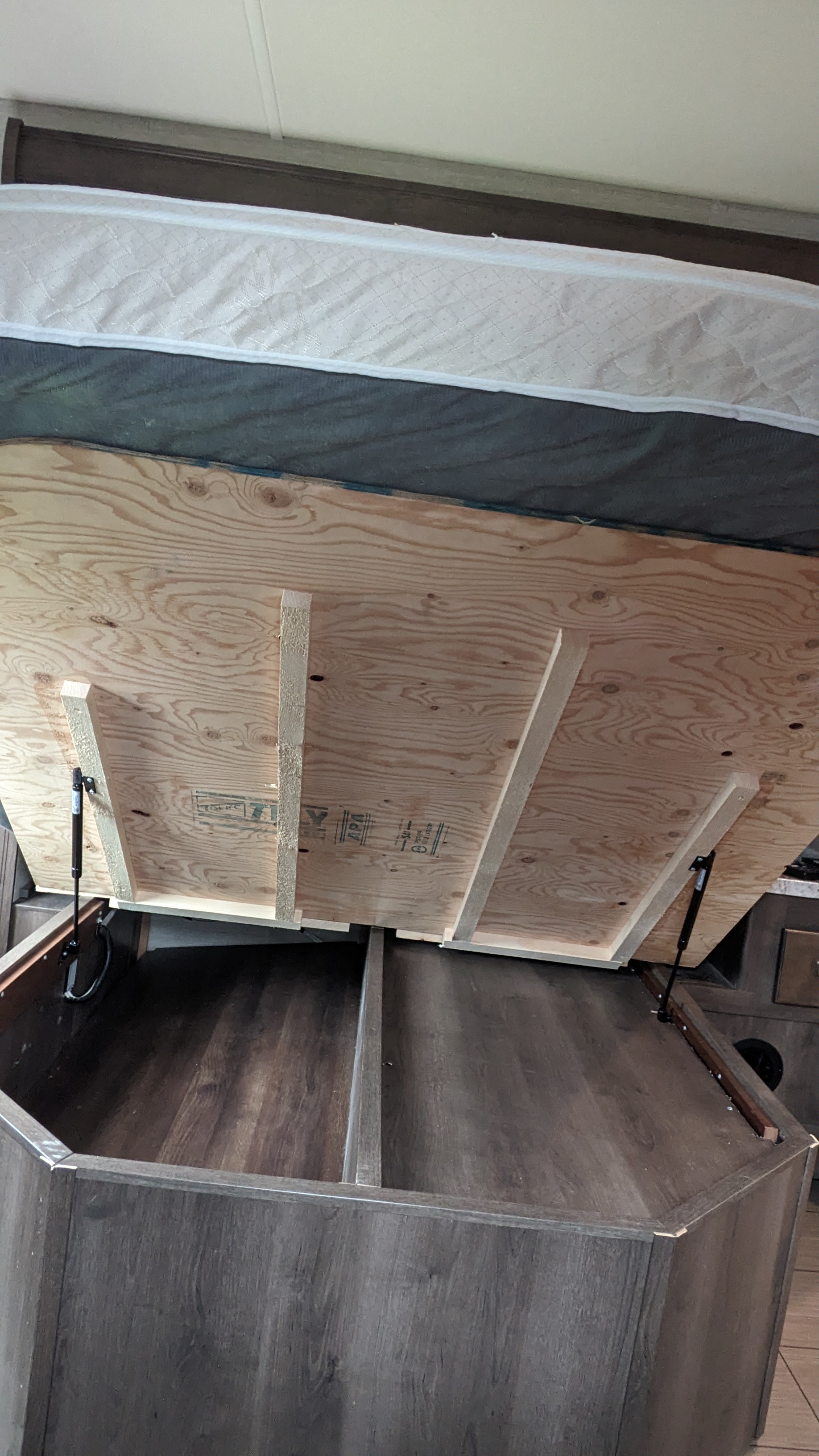 This RV rental bed features a lifted mattress, revealing convenient under-bed storage supported by gas struts and a wooden platform.. Jayco Greyhawk 2019