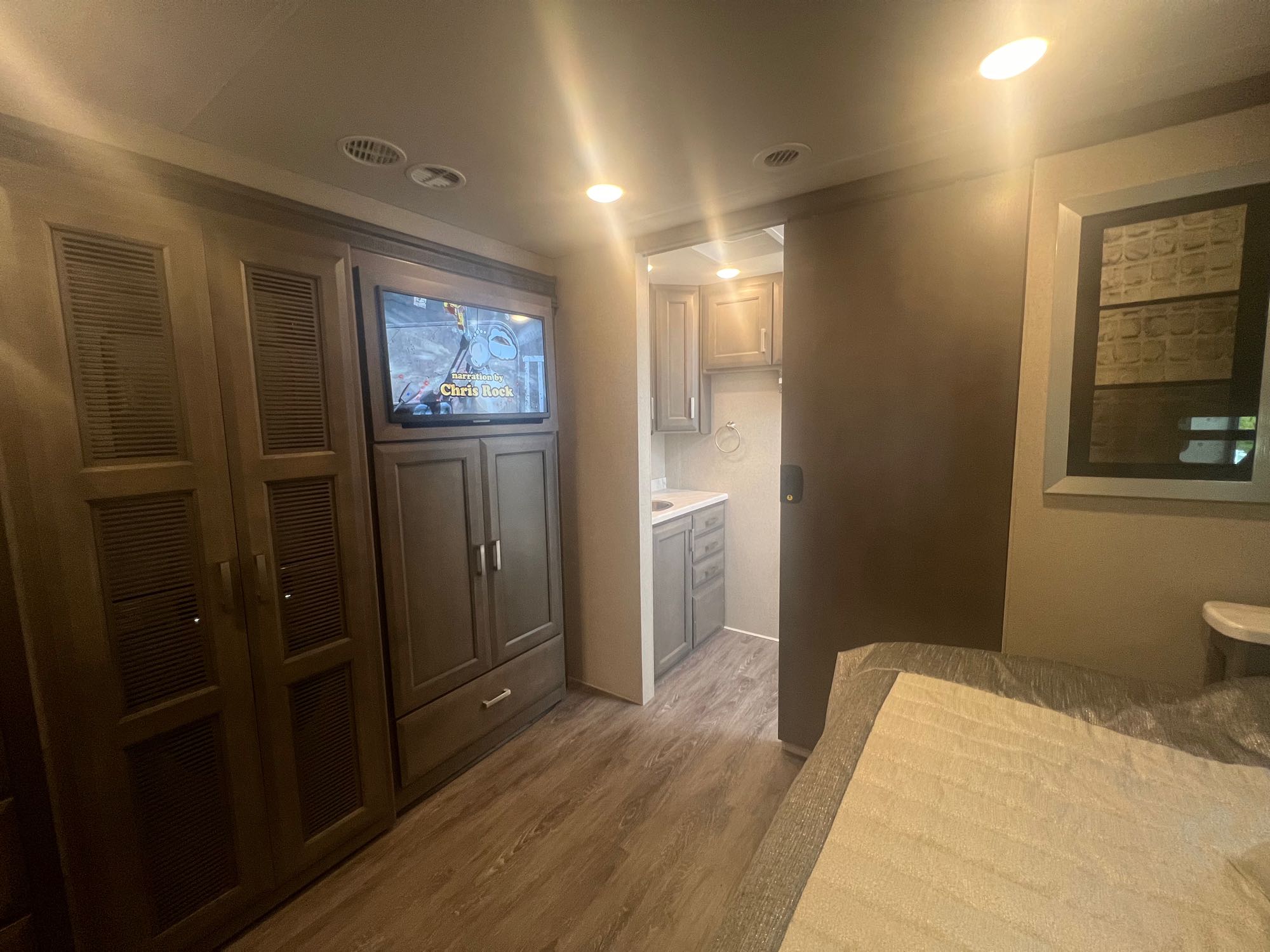 RV rental interior: bedroom with TV showing Chris Rock, open doorway to compact bathroom with sink. Wood flooring.. Thor Pasadena 2024