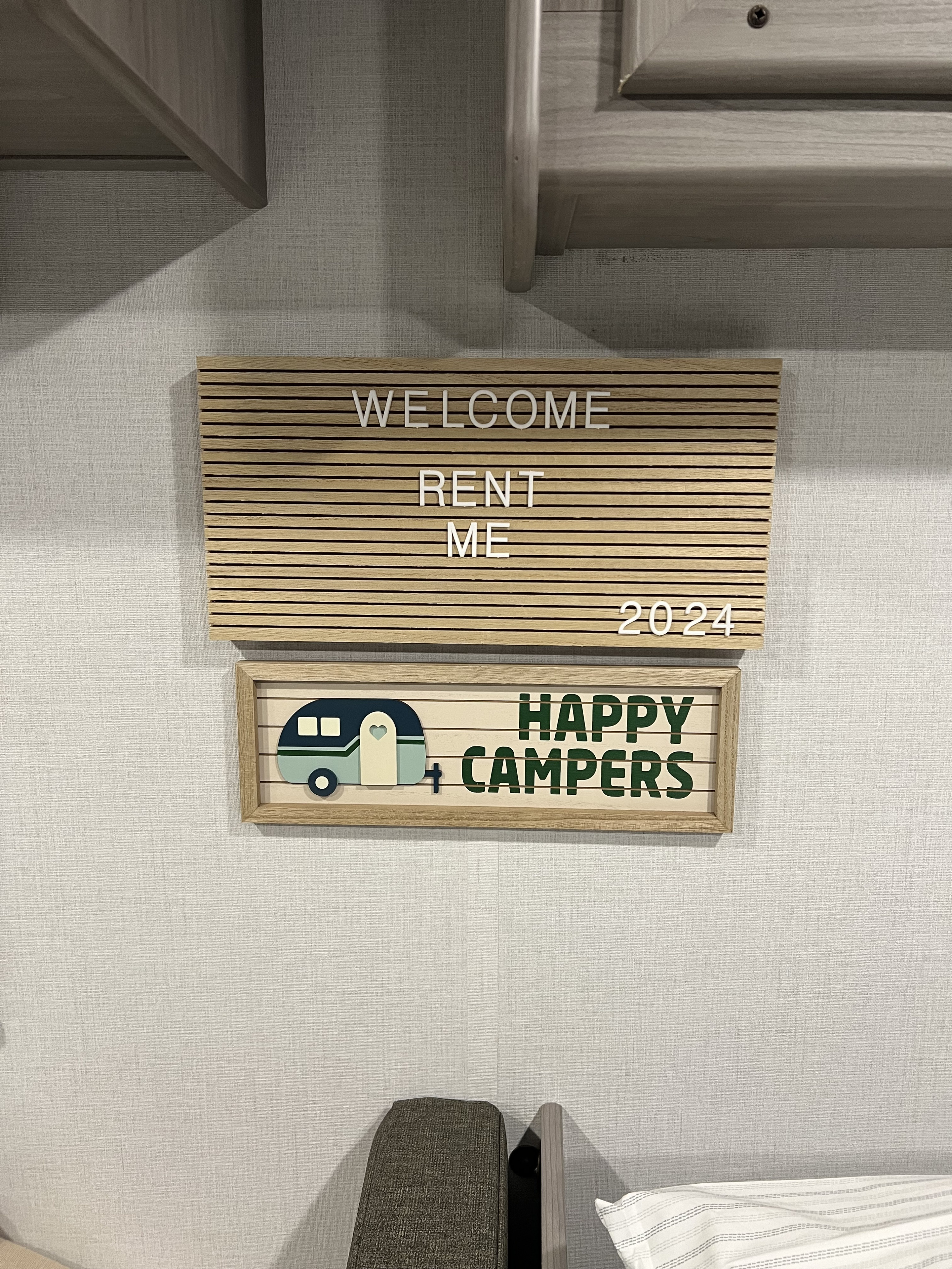 An RV rental sign reads "WELCOME RENT ME 2024" above a "HAPPY CAMPERS" plaque with a camper illustration.. Coleman Lantern 17B 2024