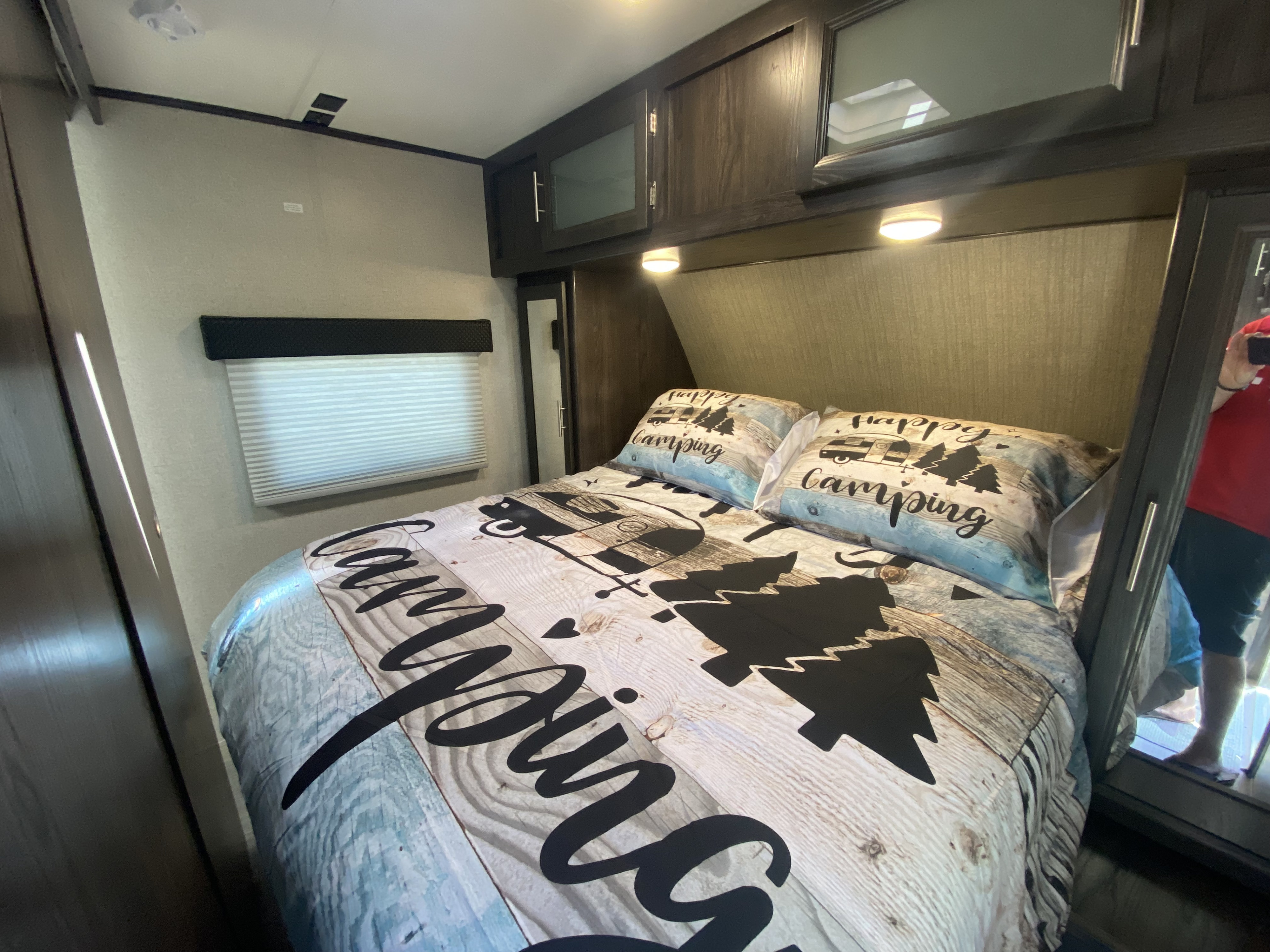 Cozy RV rental bedroom with 'Happy Camping' themed bed, overhead storage, and mirrored closet. A person is reflected in the mirror.. Dutchmen Other 2018
