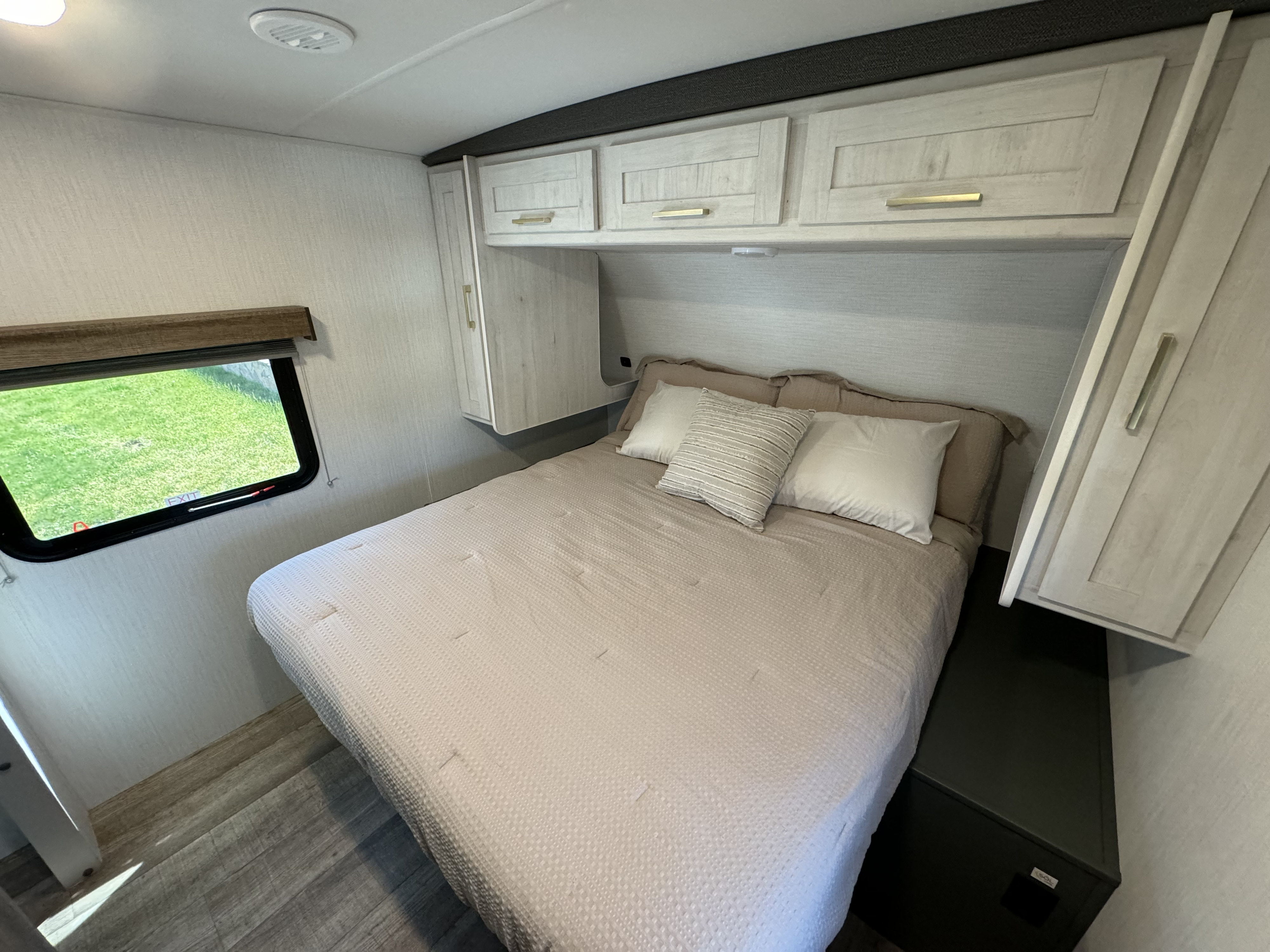 Cozy RV rental bedroom featuring a comfortable bed, overhead cabinets, window view, and wood-look flooring.. Heartland RVs ID260RB 2024