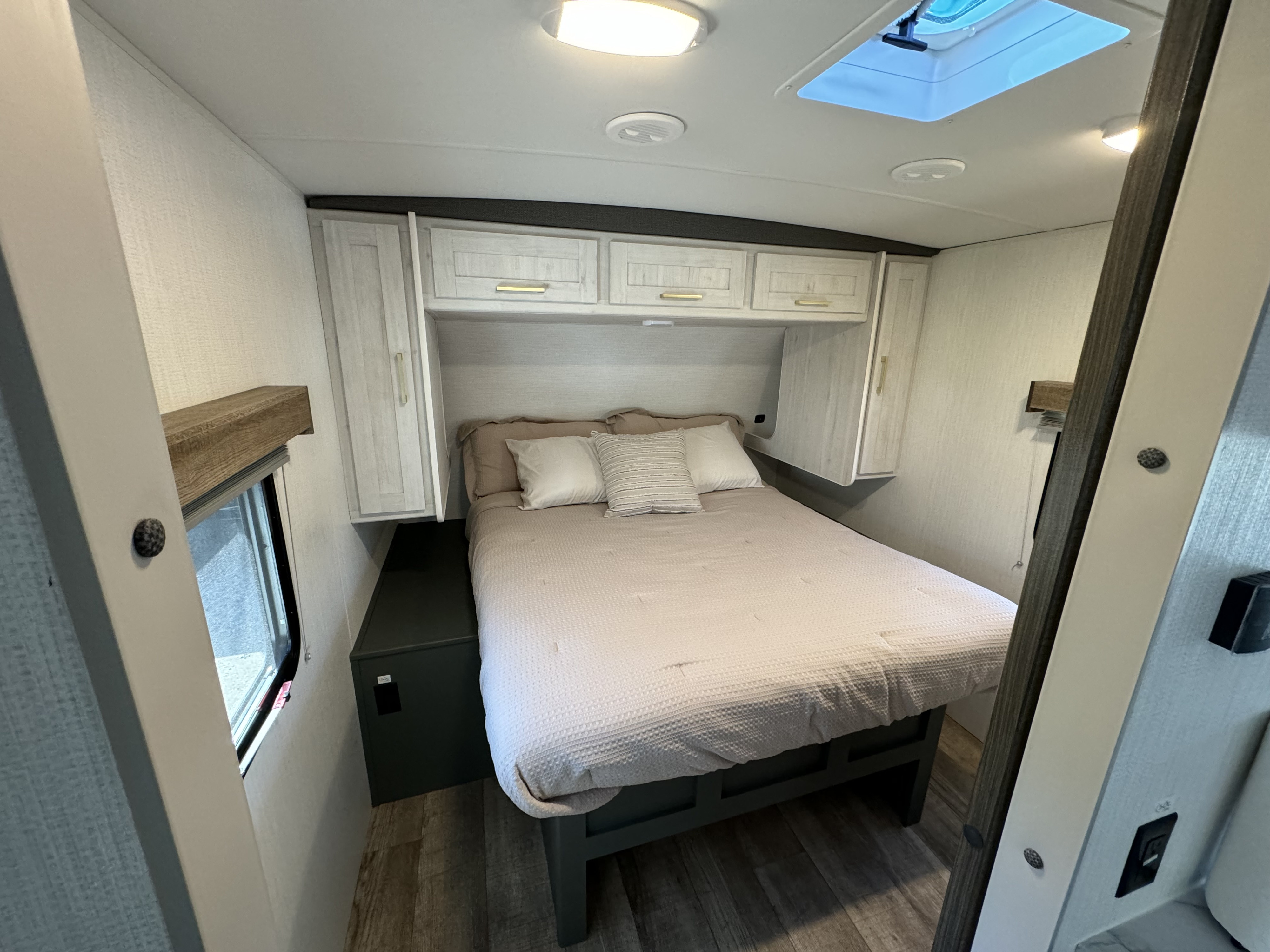 This cozy RV rental bedroom features a comfortable bed, overhead cabinets, and a window, with light walls and wood floors.. Heartland RVs ID260RB 2024