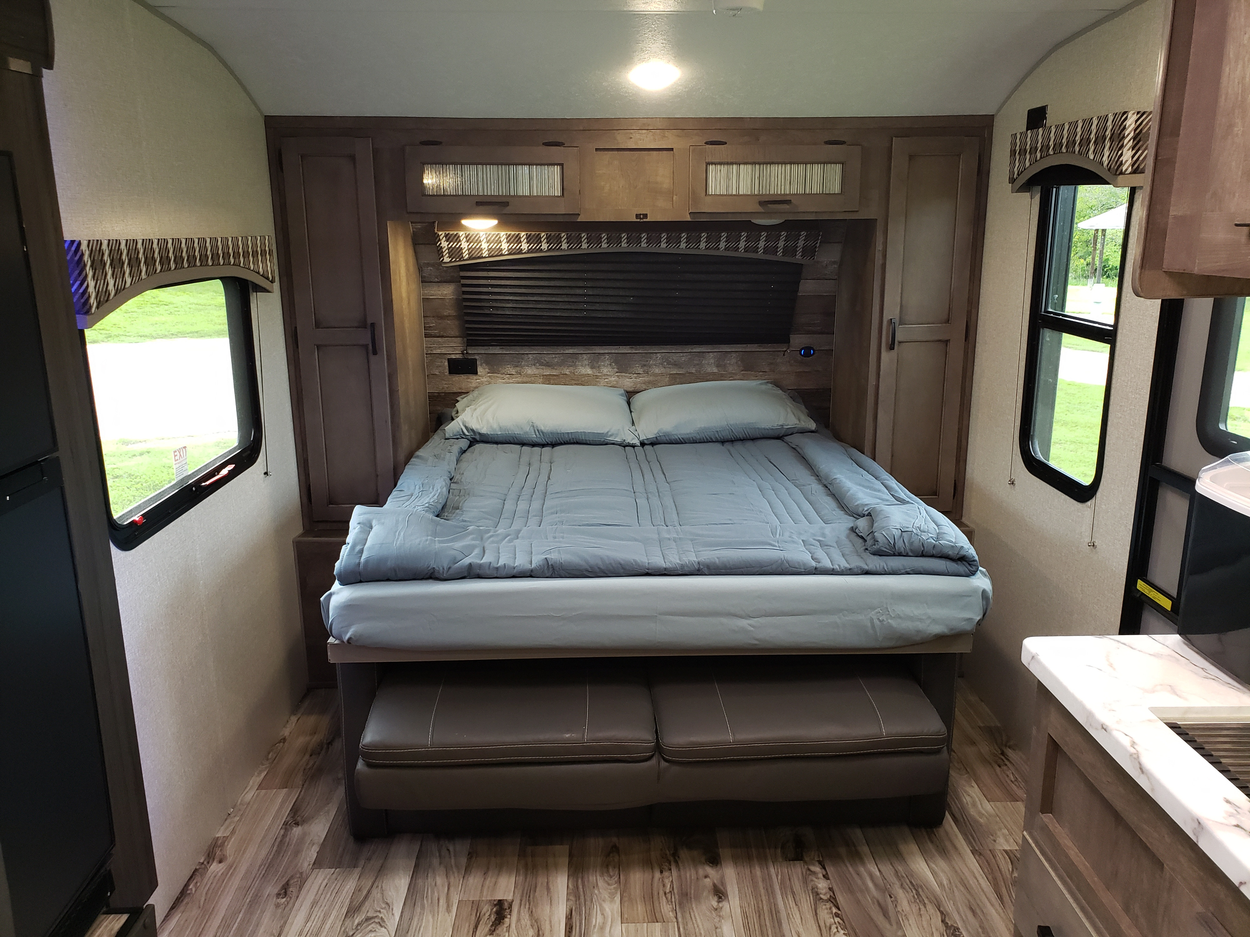 Cozy RV rental bedroom with a neatly made bed, light blue sheets, wood-look cabinetry, and windows overlooking green scenery.. Venture Sonic SN211VDB 2022