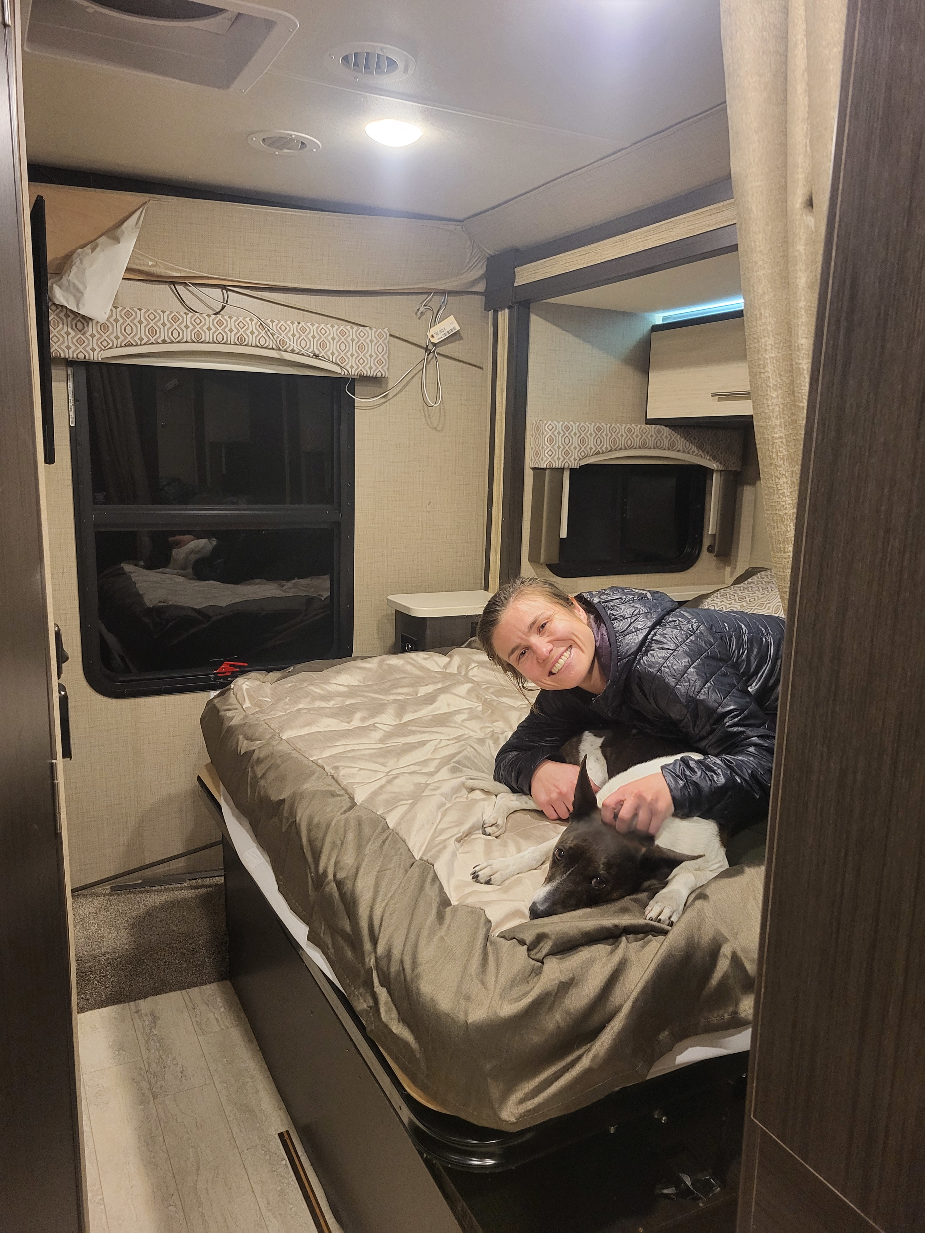 Smiling woman and dog relax on bed in a cozy RV rental bedroom, showing windows, bed, and RV interior details.. Thor Vegas 2020