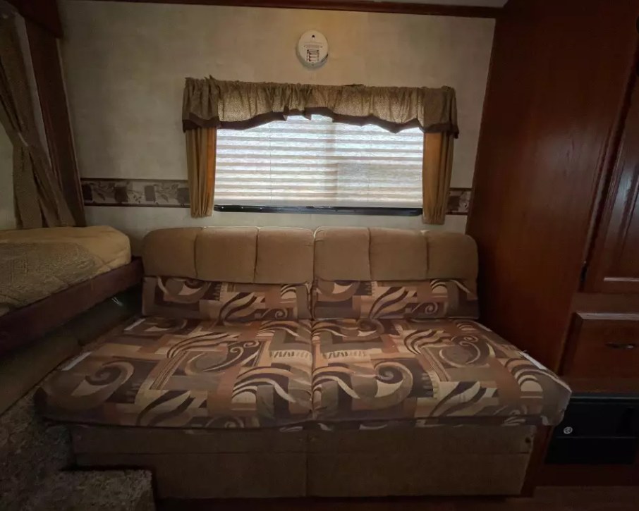 RV rental interior: patterned sofa, window with blinds, and a visible bed. Wood accents add warmth to this mobile living space.. Keystone RV Outback 2010