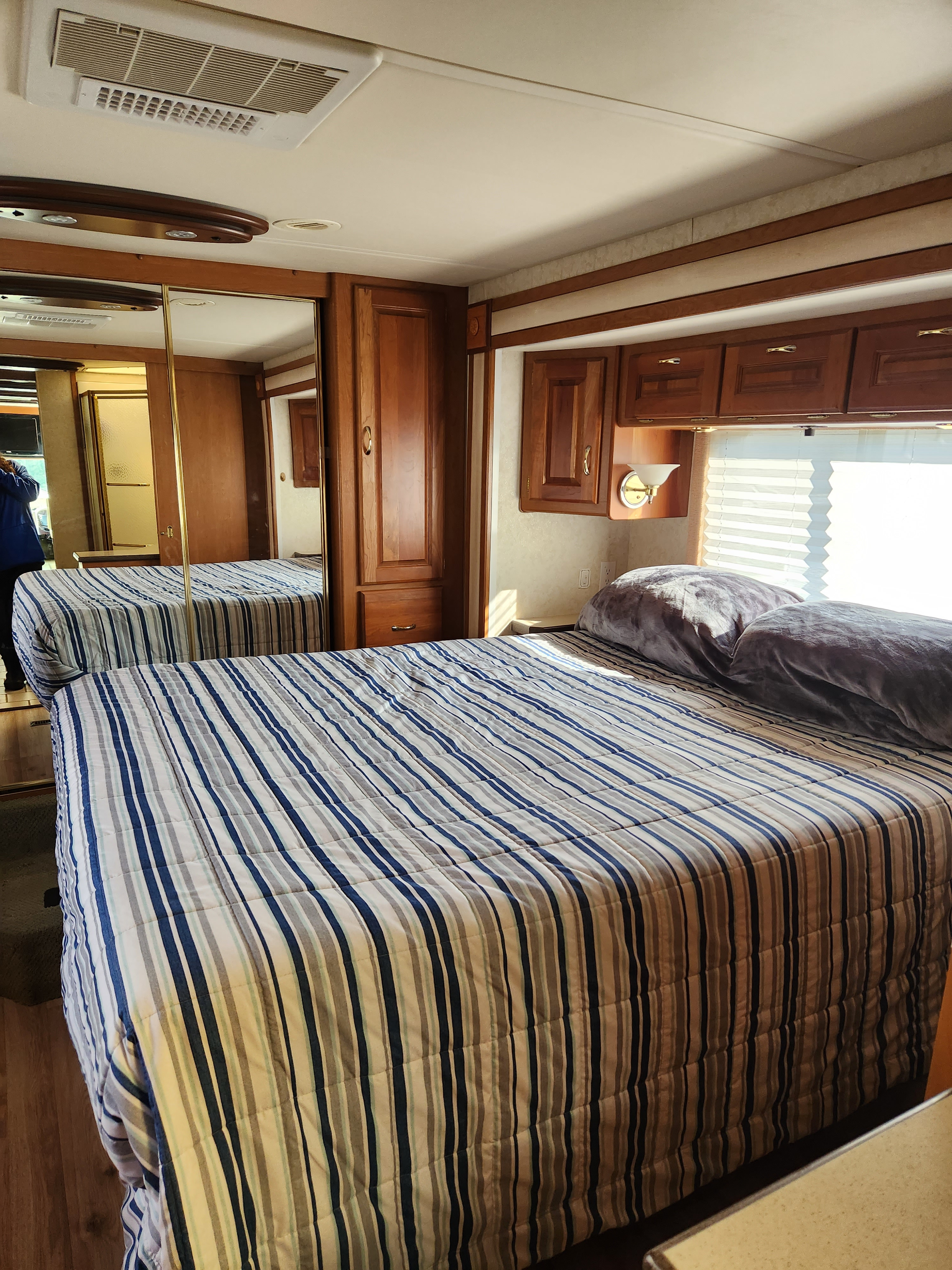 Step inside this cozy RV rental bedroom. Features a striped bed, mirrored wardrobe, wood cabinets, and bright window.. Holiday Rambler Endeavor 2003