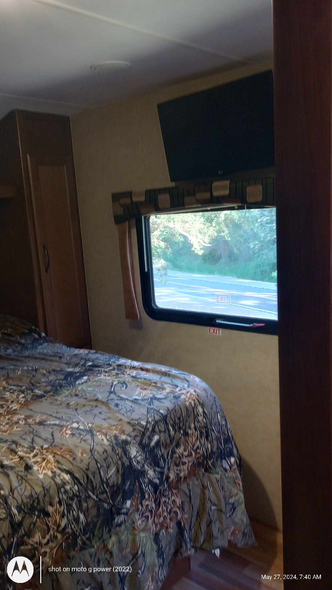 An RV rental bedroom features a bed with camo bedding, a wall-mounted TV, and an emergency exit window overlooking a road.. Pacific Coachworks Northland 2016