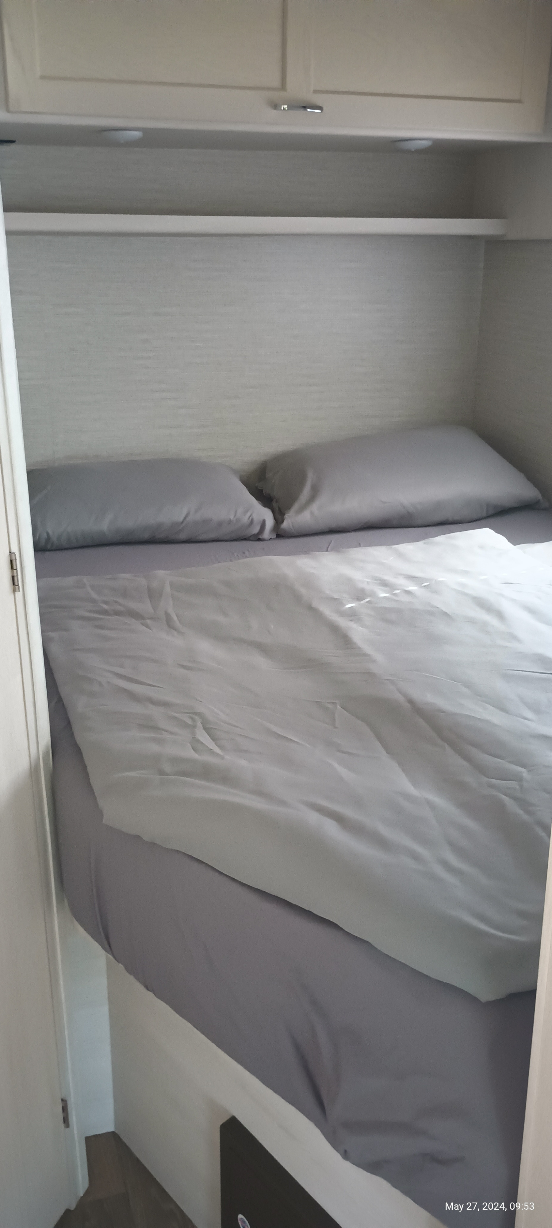 RV rental bed with grey pillows and duvet under overhead cabinets and shelf, a compact sleeping area.. Winnebago Outlook 2021