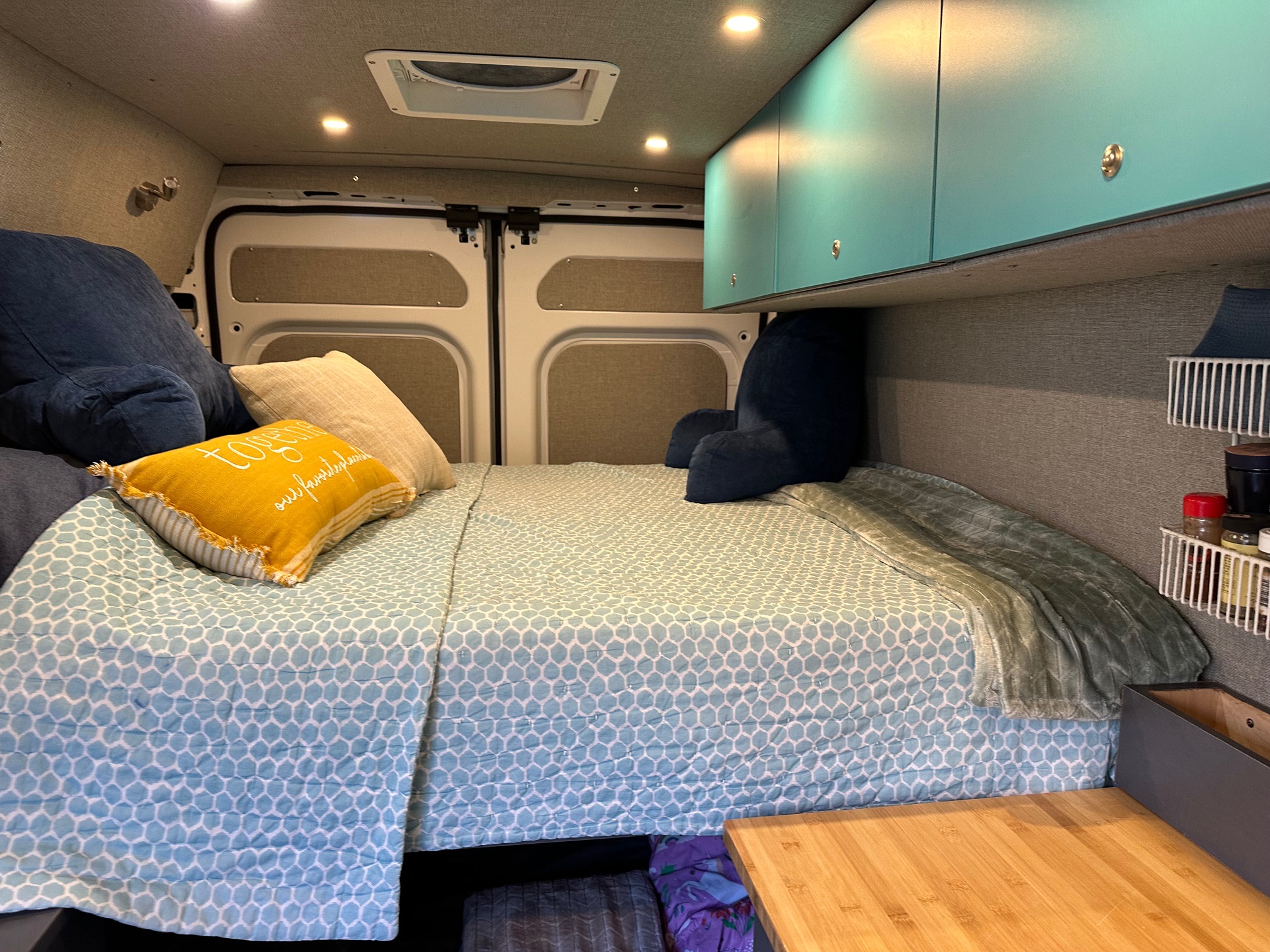 Cozy RV rental interior: a comfy bed with blue bedding, teal overhead cabinets, and compact storage for travel.. RAM ProMaster 2023