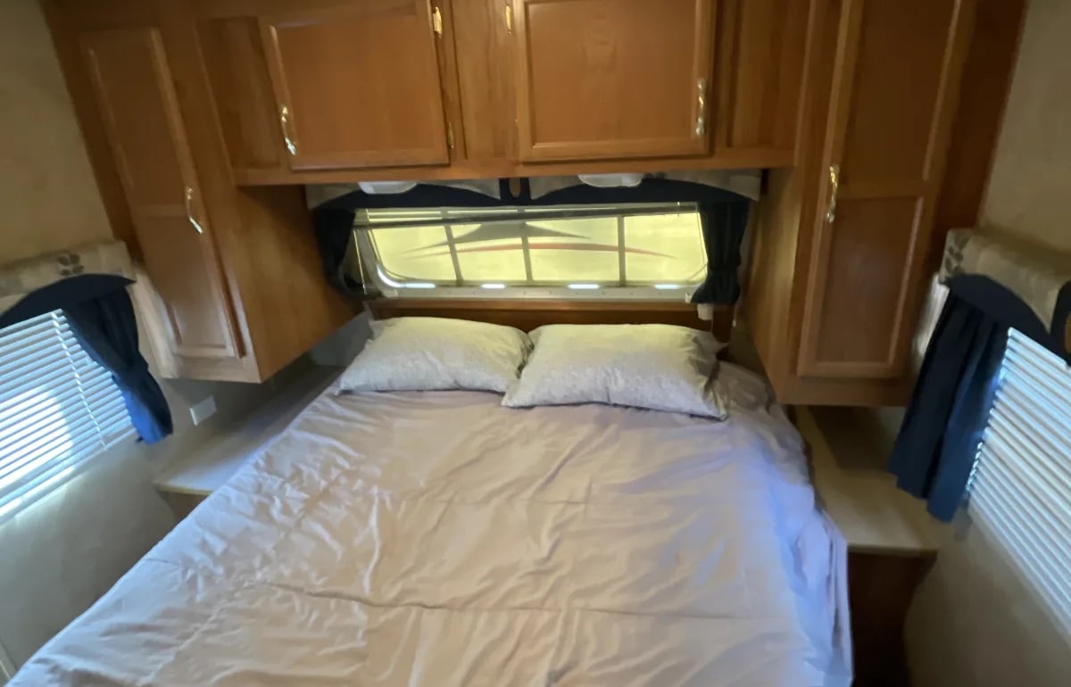 Compact RV rental bedroom with a bed, pillows, overhead cabinets, and windows on three sides for natural light.. Jayco Jay Flight 2006
