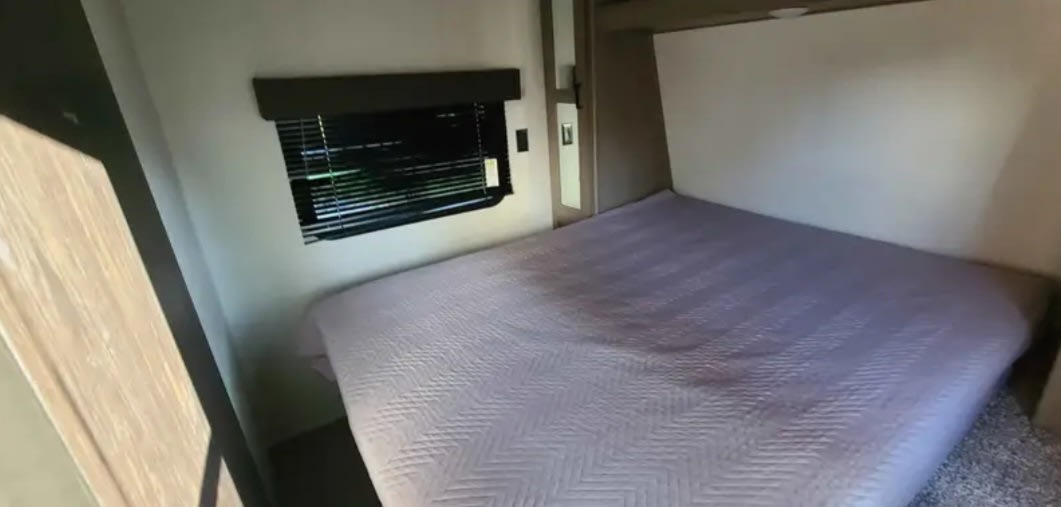 RV rental bedroom: a tidy bed with a light purple quilt, window with blinds, and built-in bedside storage.. Keystone RV Hideout LHS 2020