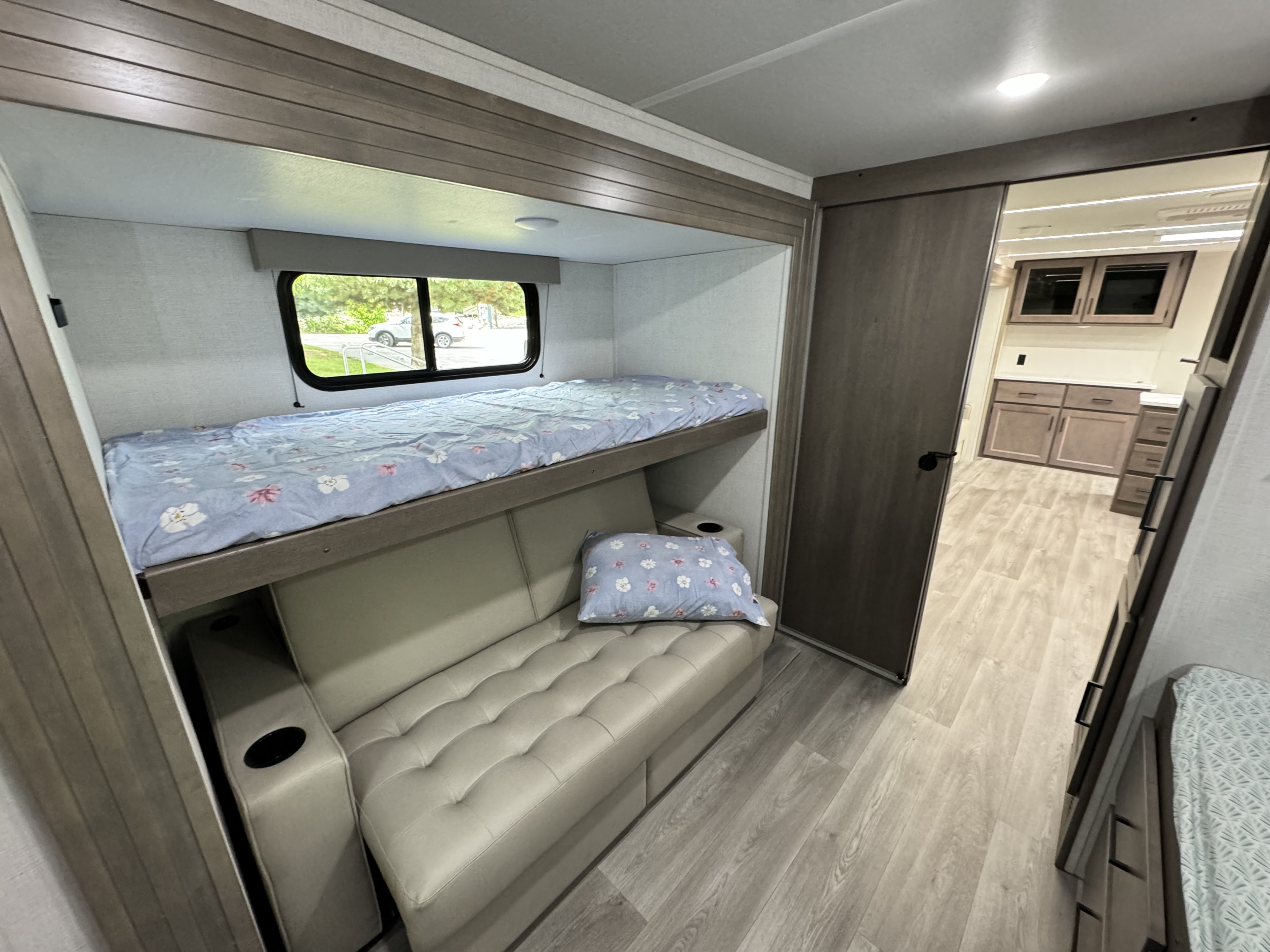 RV rental interior: cozy bunk beds with floral bedding, a comfortable tufted sofa, and a modern kitchen beyond.. Grand Design Transcend Xplor 2024