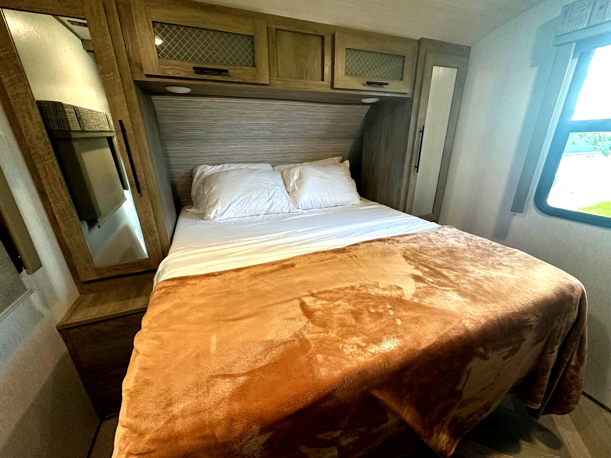 Inside a cozy RV rental: a comfortable bed with a brown blanket, overhead cabinets, and a window to the outdoors.. Forest River Wildwood Heritage Glen HL 2022
