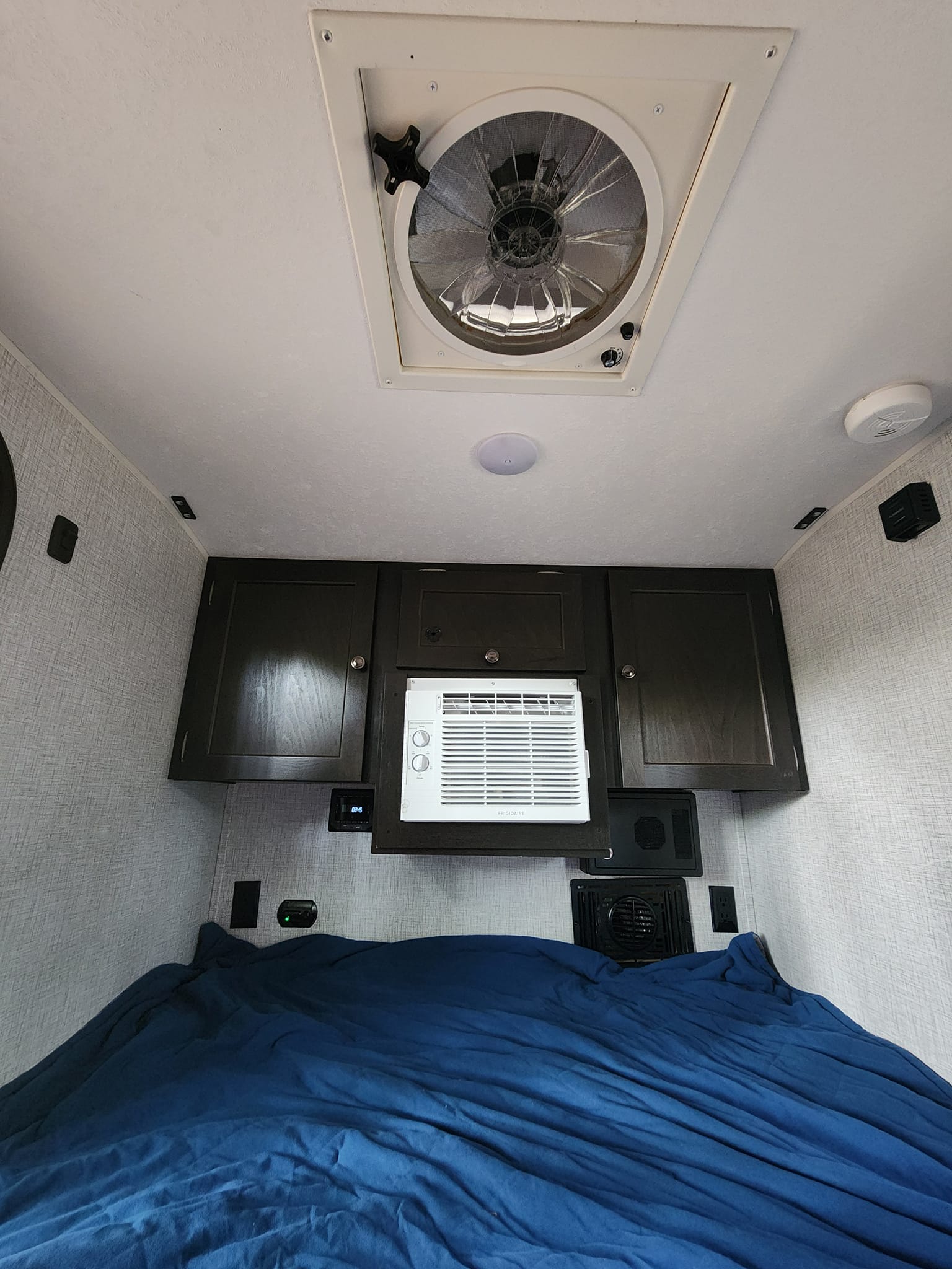 This RV rental bedroom features a comfy bed with blue sheets, a wall-mounted AC unit, dark wood cabinets, and a ceiling fan.. Braxton Creek Free Solo 2021