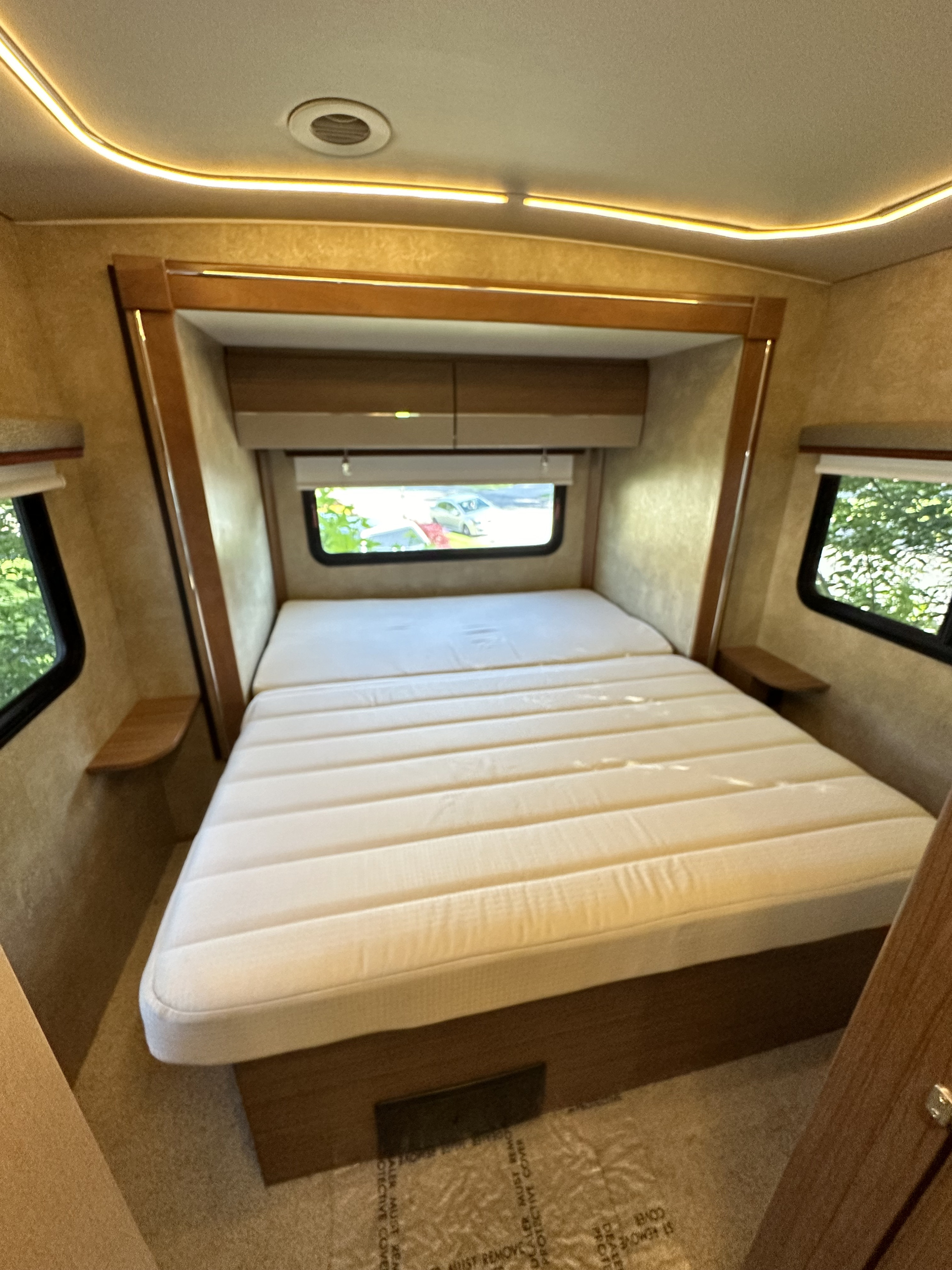 Inside an RV rental bedroom featuring a large bed, windows, overhead cabinets, and warm ambient ceiling lighting.. Winnebago View 2017
