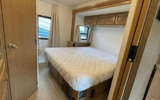 This RV rental features a cozy bedroom with a comfortable bed, overhead cabinets, and a window for natural light.. Thor Windsport 2019