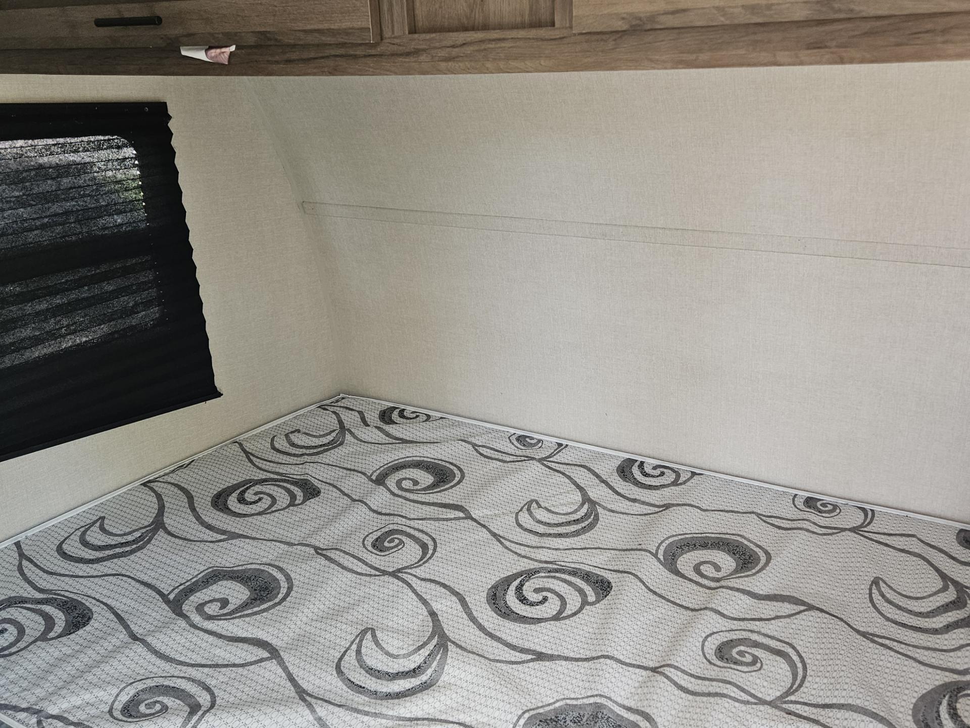 RV rental interior: light-walled sleeping area with a patterned mattress, black window blind, and wood cabinets overhead.. Avenger LT 2021