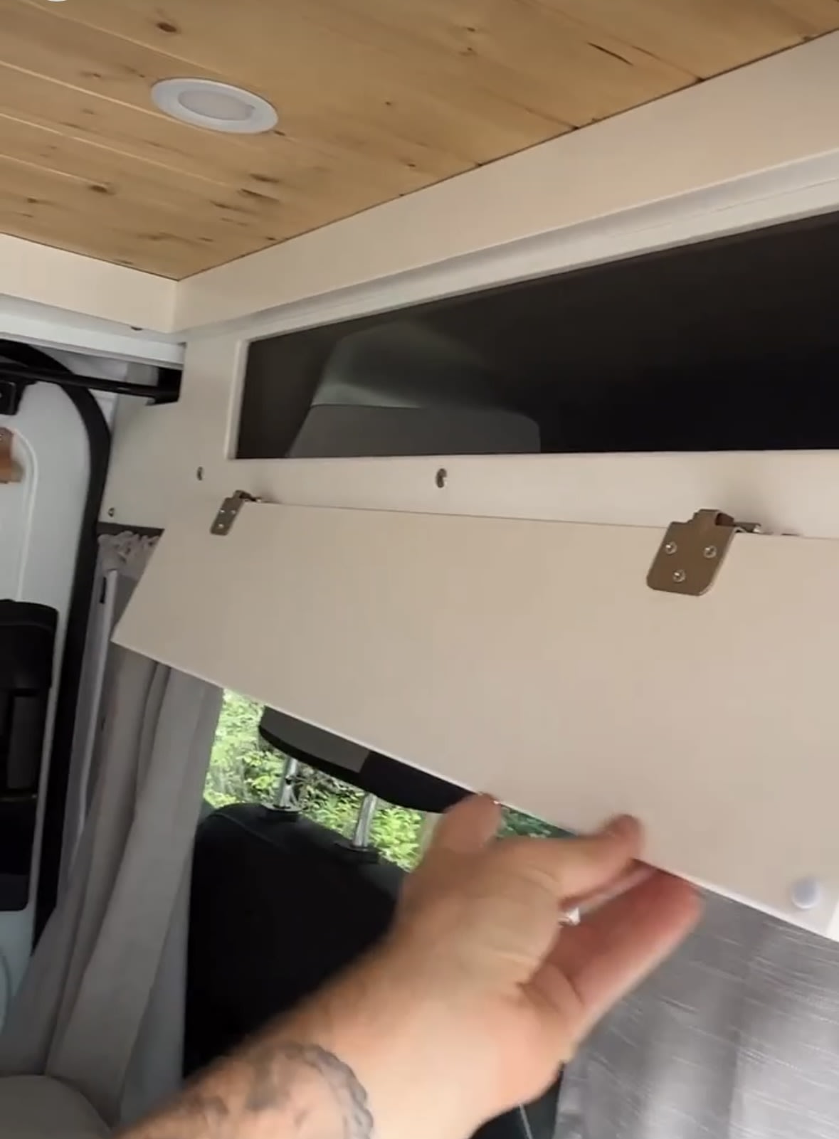Inside an RV rental, a hand opens a white overhead storage cabinet with a wooden ceiling and window view visible.. Ford Transit Custom 2019
