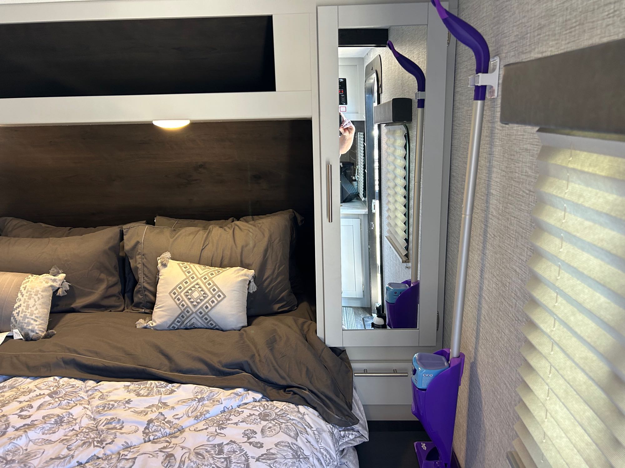 Inside an rv rental, a compact bedroom features a made bed, overhead storage, and a wall-mounted Swiffer next to a mirror reflecting the interior.. Keystone RV Bullet Ultra Lite 2021